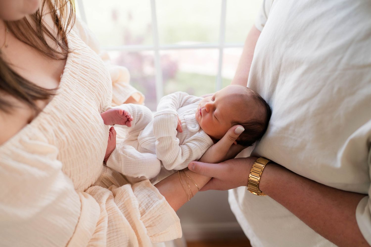 Two parents cradle their sleeping newborn baby together, with mother in cream dress and father wearing gold watch.
