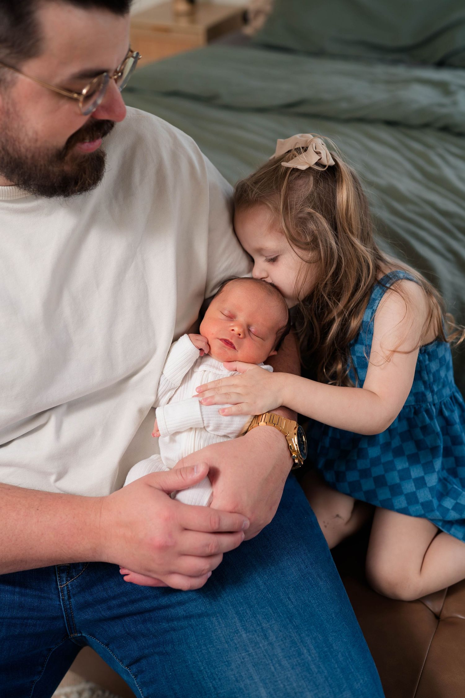 Father holds swaddled newborn while young girl in blue dress kisses baby's head during family photo session.