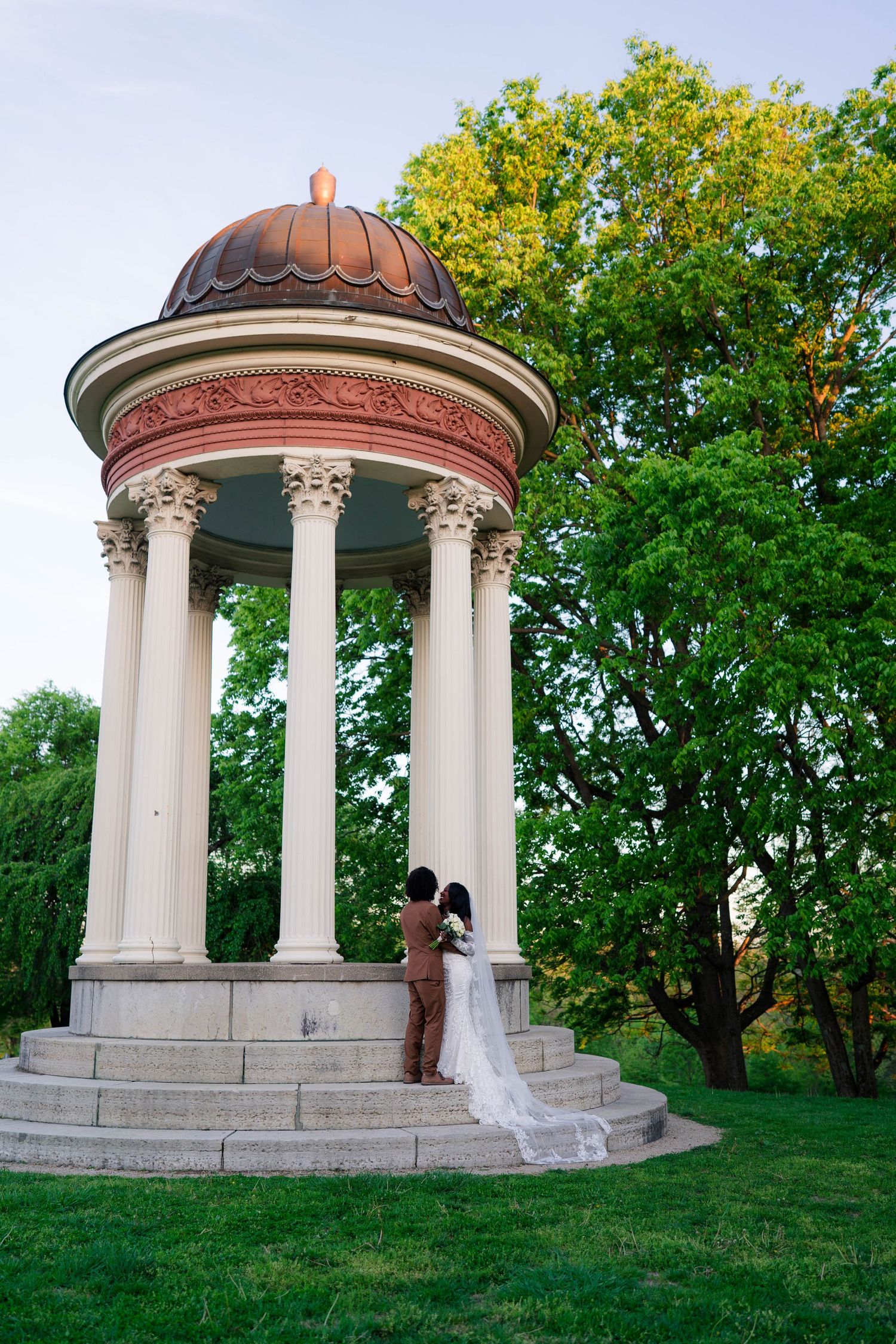 Bride and groom embrace under a classical rotunda gazebo with white columns and copper dome in a lush green park.