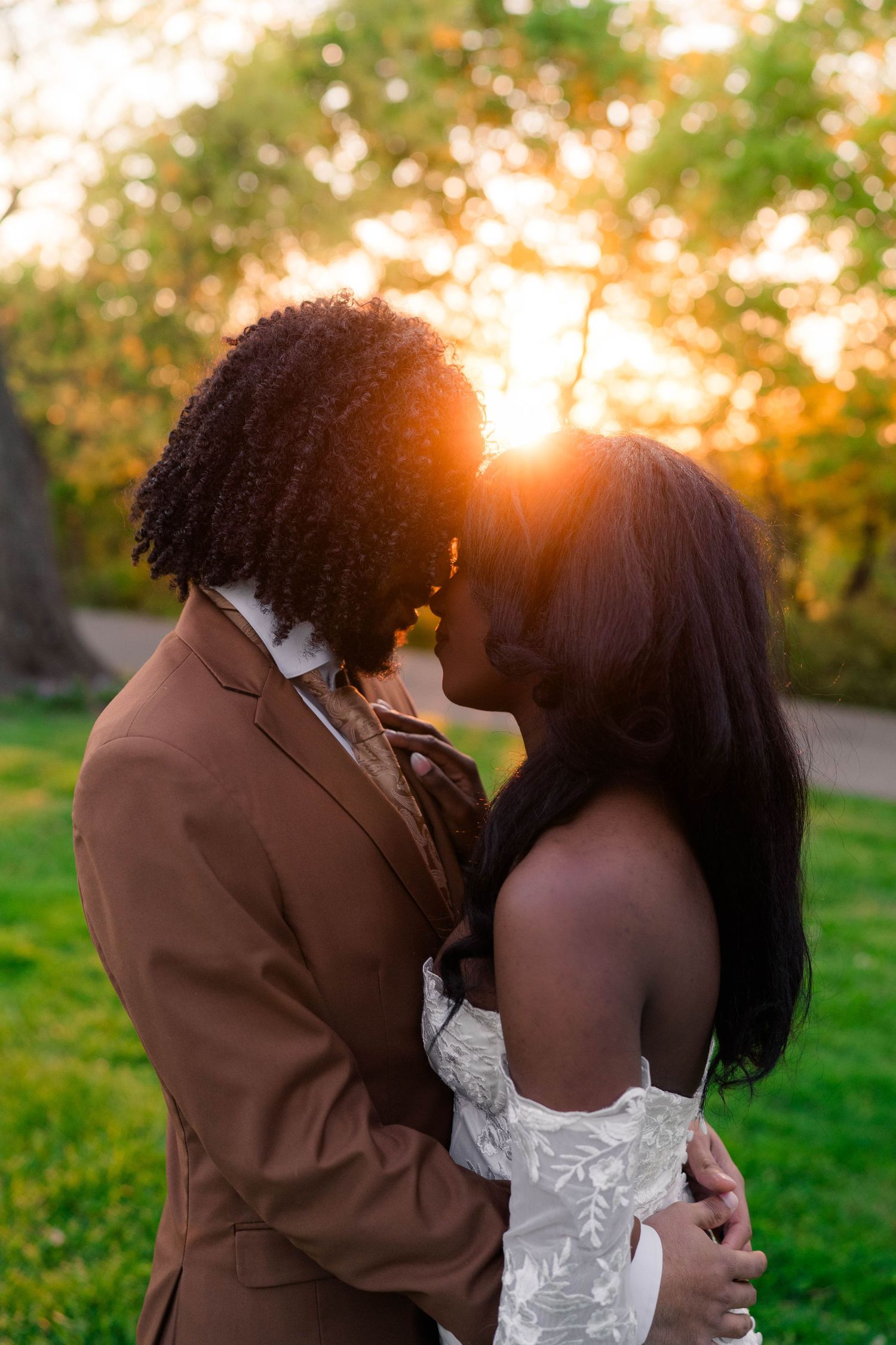 Couple sharing a romantic kiss at golden hour sunset, groom in brown suit and bride in white lace dress outdoors.