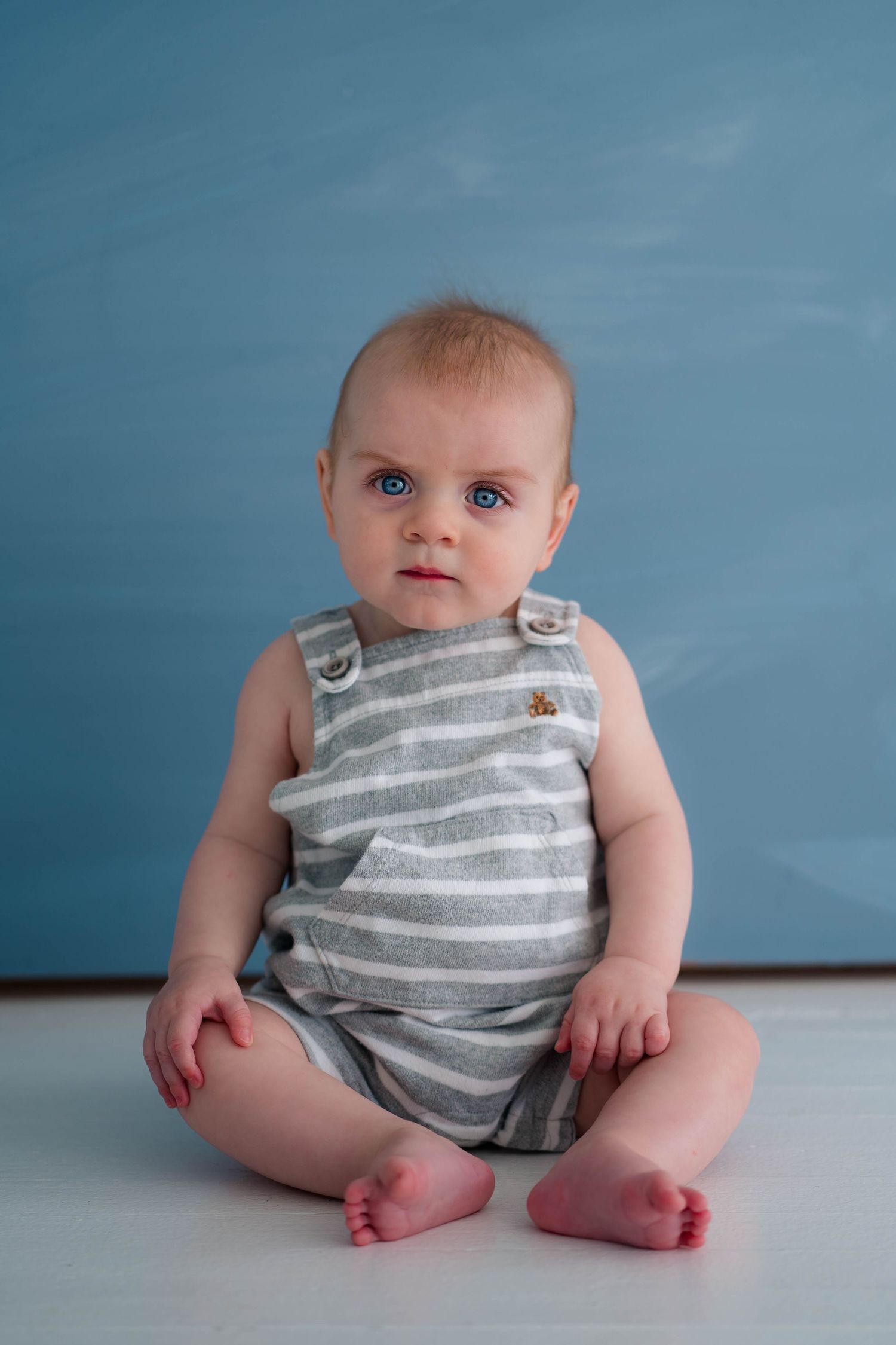 Cute baby with blue eyes sitting on white floor wearing gray striped romper against blue background.