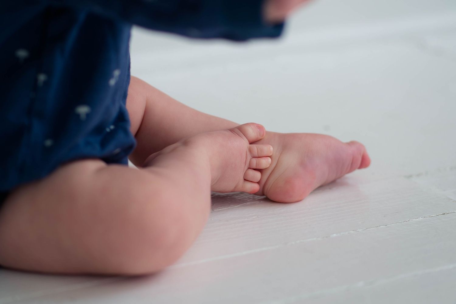 Close-up of baby's bare feet and legs in a navy blue outfit on white floor.