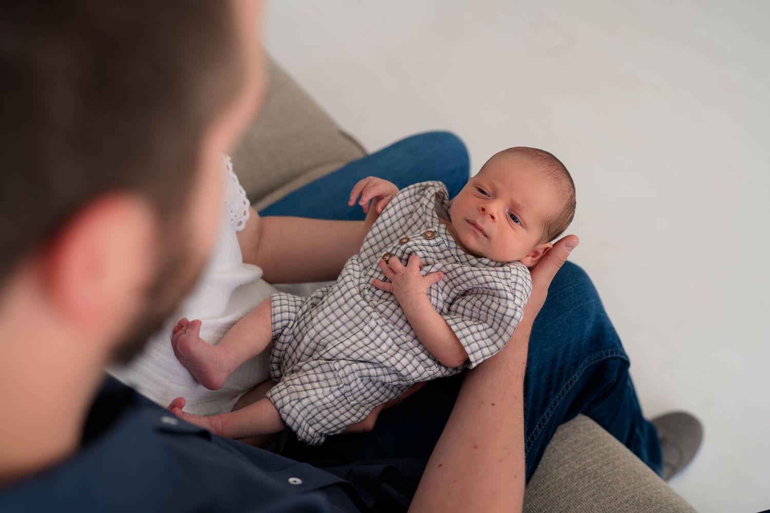 Father holding peaceful newborn baby in plaid outfit on cozy couch from overhead view.