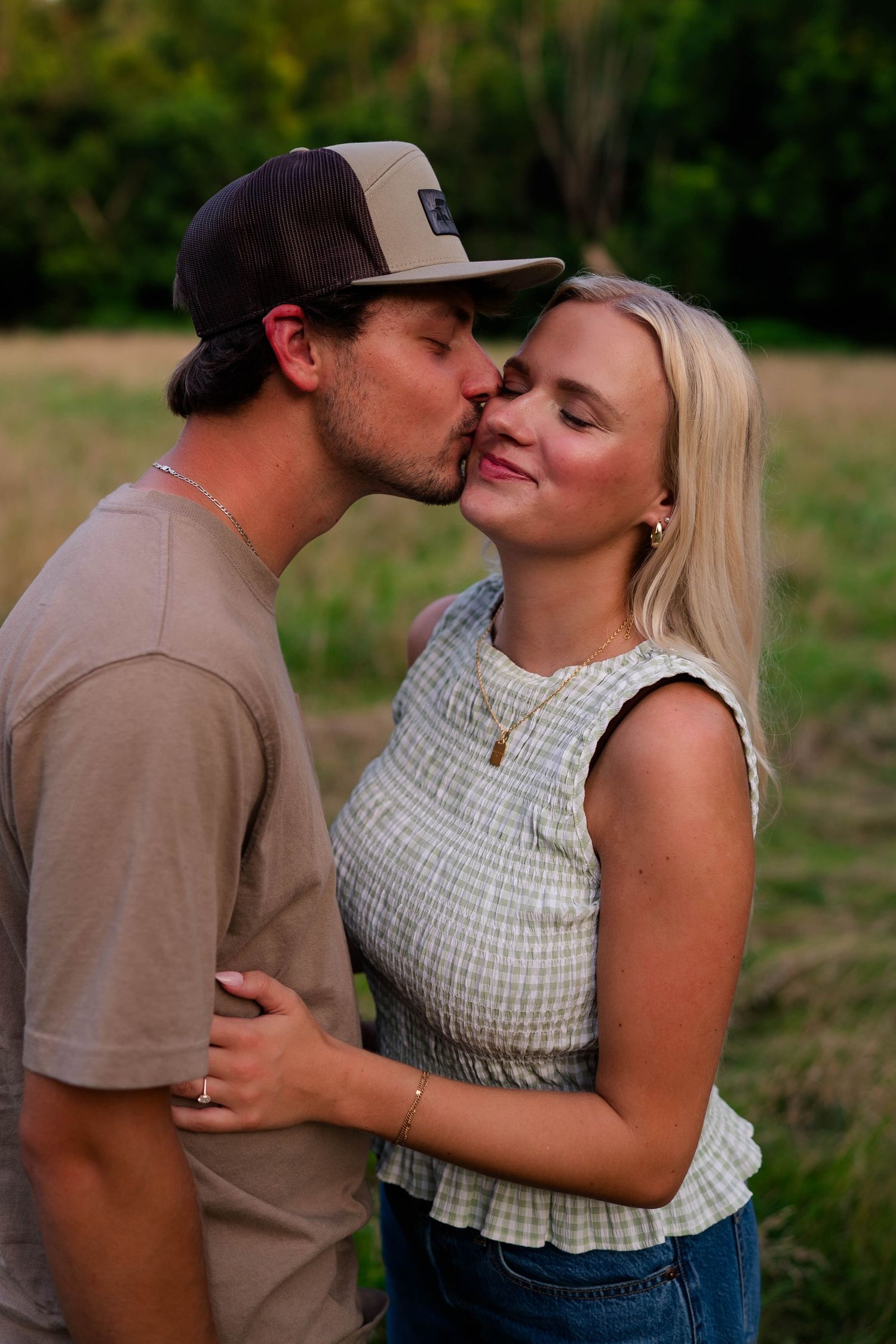 A young couple shares a tender kiss in a lush green field, the man wearing a brown cap and t-shirt.