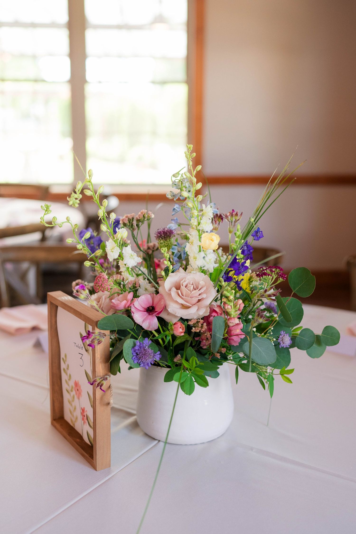 Colorful wildflower centerpiece with pink roses in white pot beside wooden frame on table.