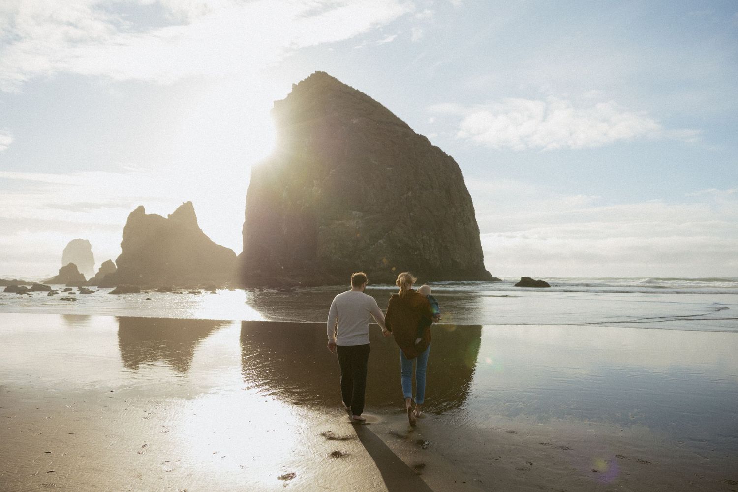 Two people strolling along a reflective beach toward Haystack Rock at golden hour, waves gently lapping the shore.