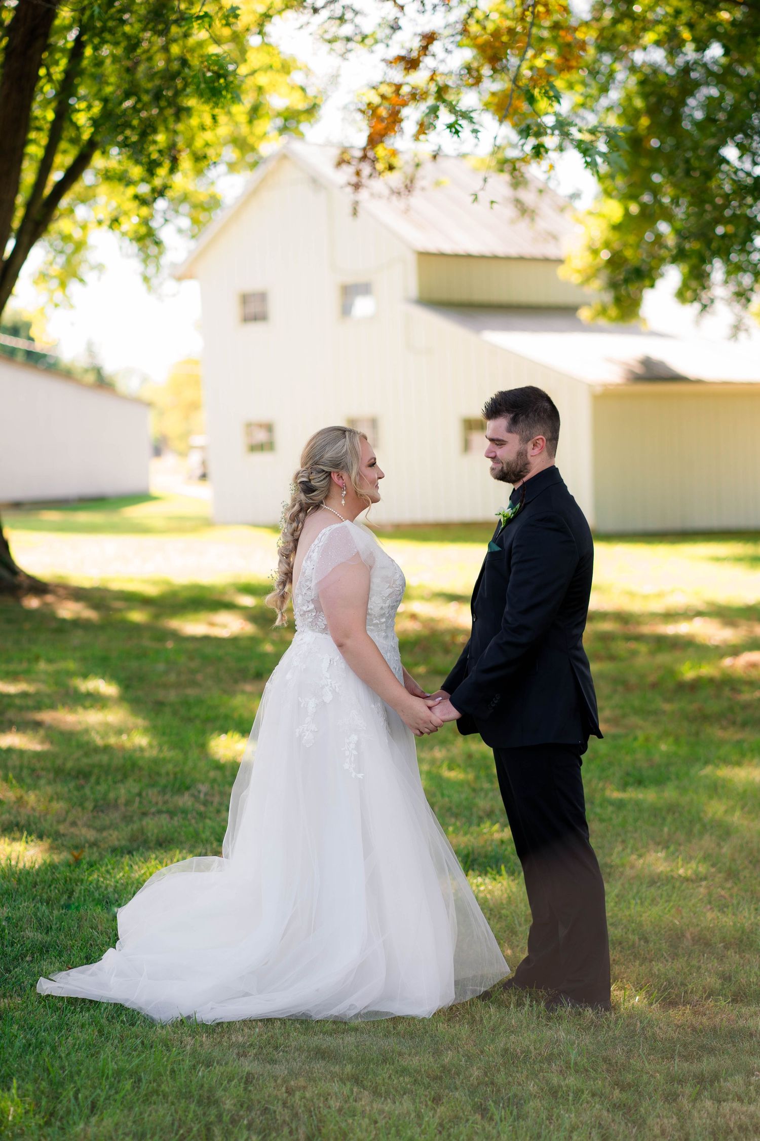 Bride and groom hold hands facing each other on a green lawn beside a white barn.