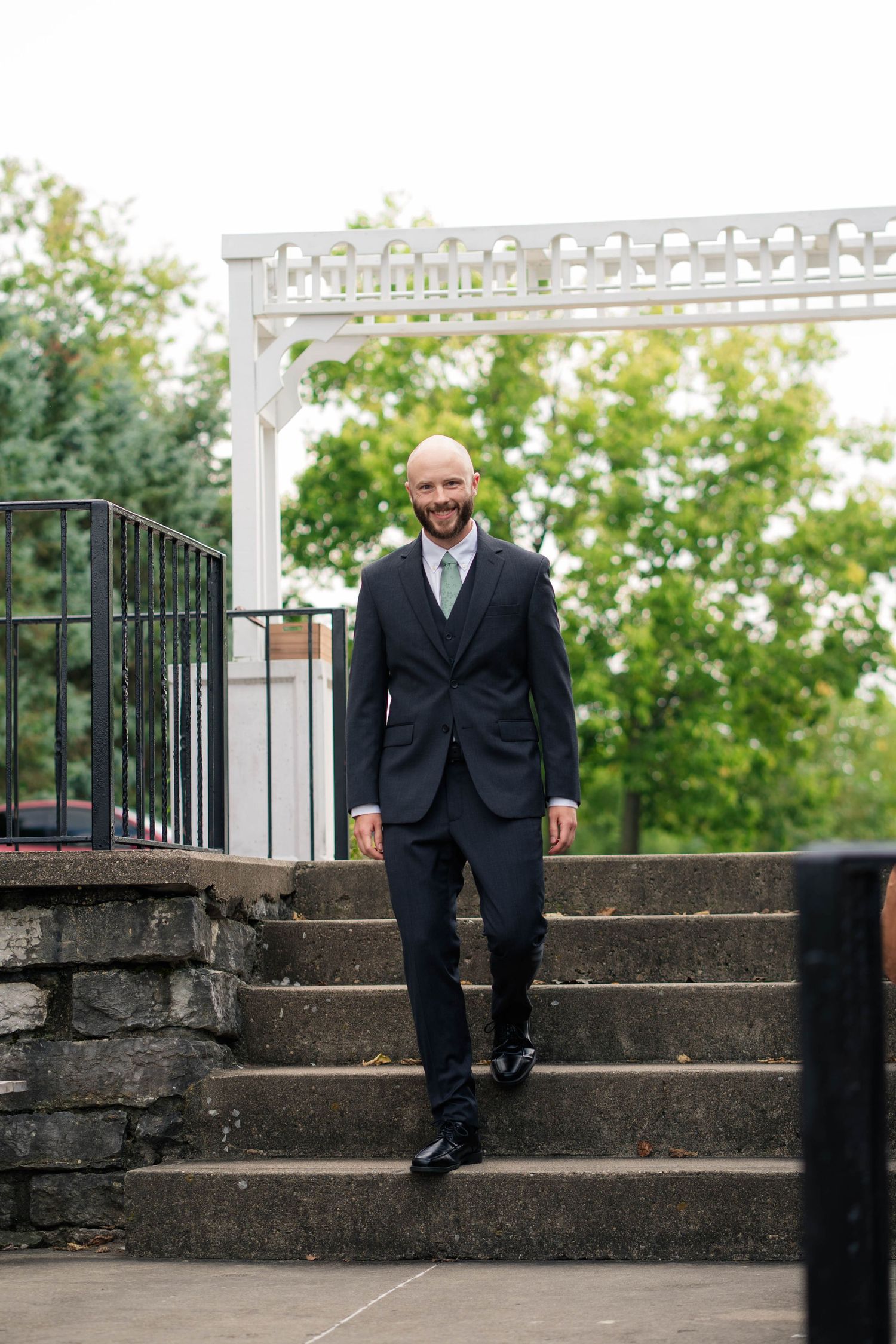 Smiling bald groom in dark suit with green tie descending stone steps at outdoor wedding venue.