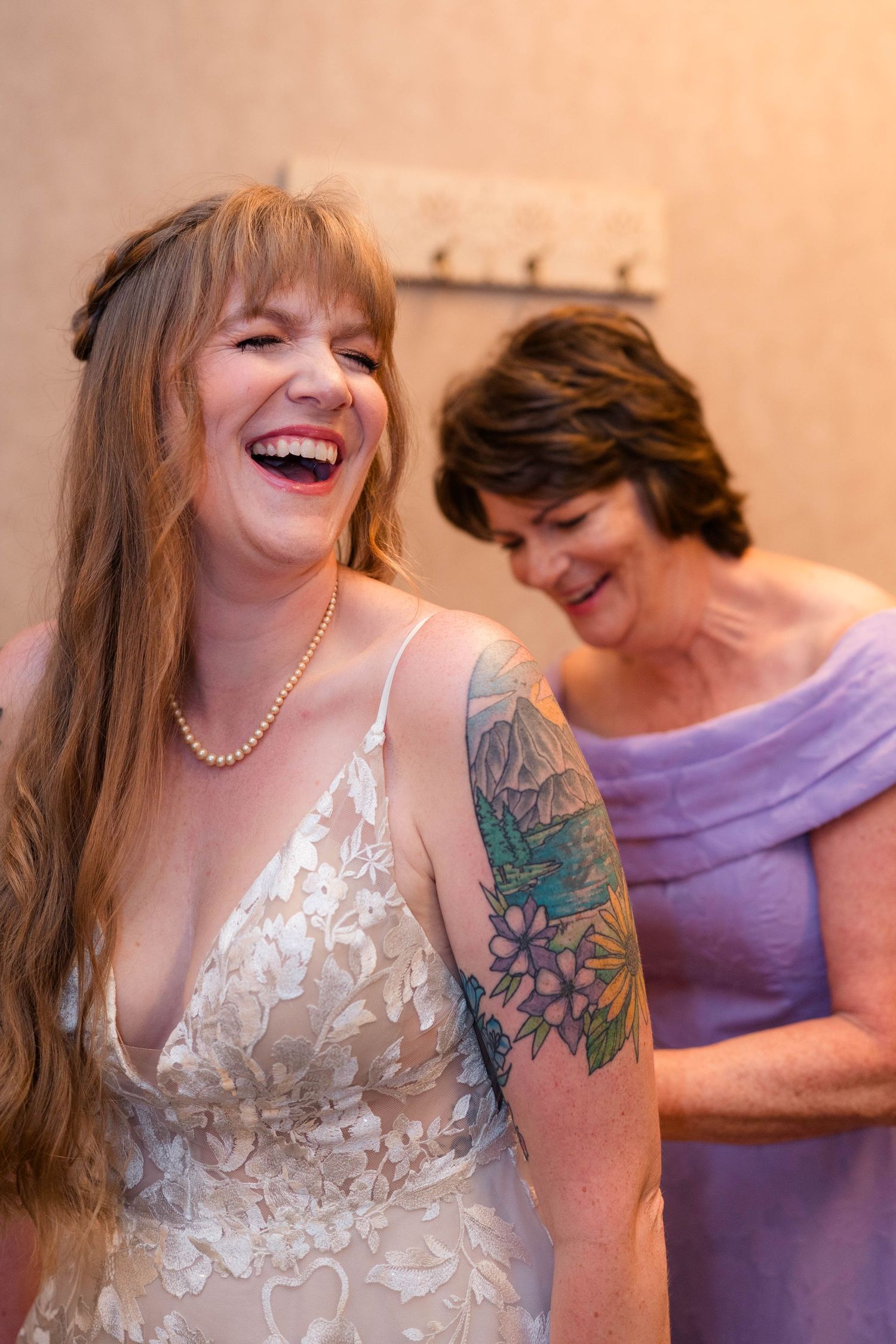 Laughing bride in lace wedding dress with floral arm tattoo and pearl necklace getting ready with woman in lavender dress.