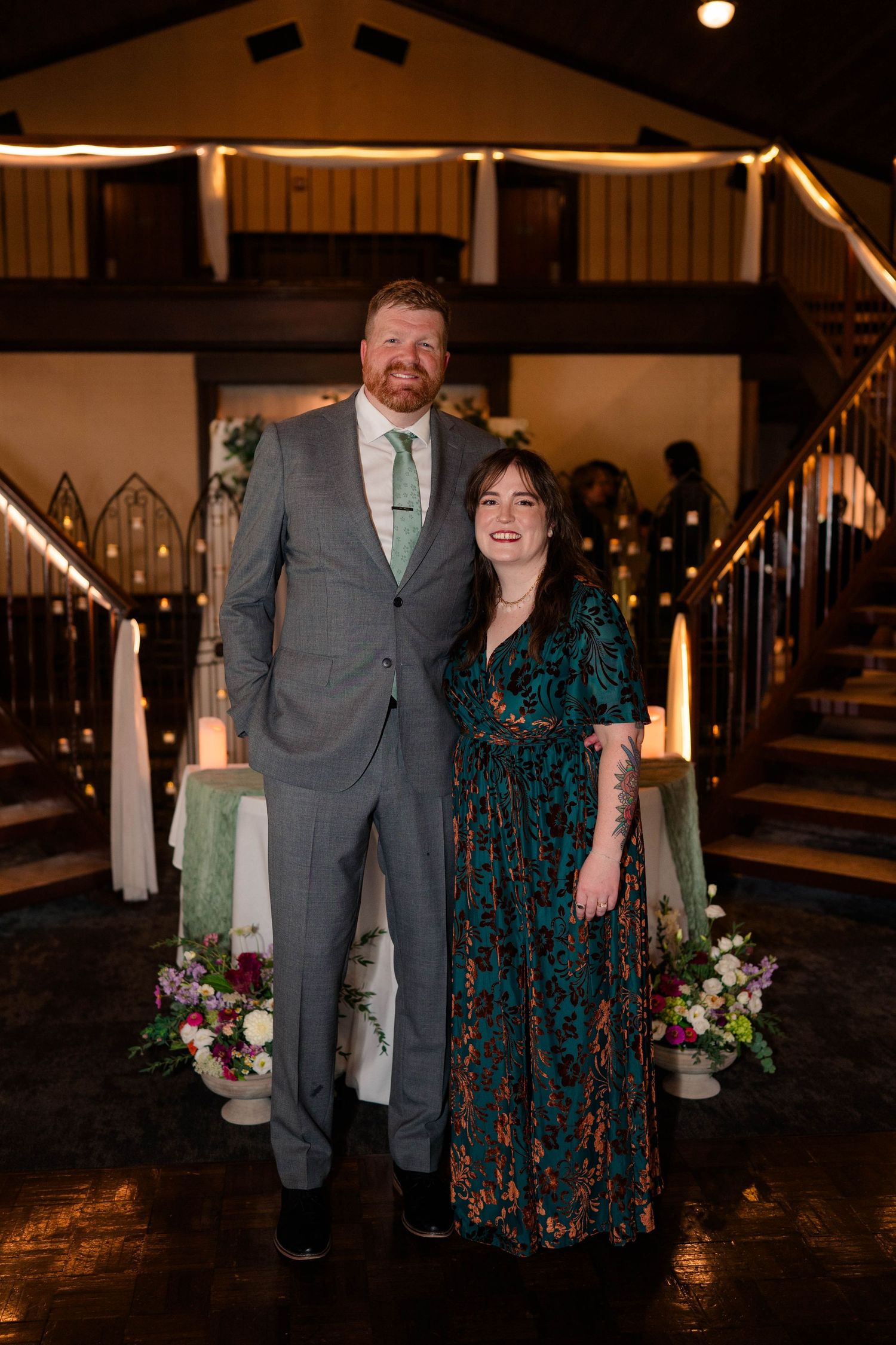 Couple posing together at a wedding venue, man in gray suit and woman in green floral dress.