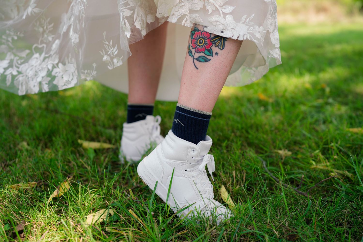Close-up of bride's legs showing ankle tattoo and white sneakers under lace gown.
