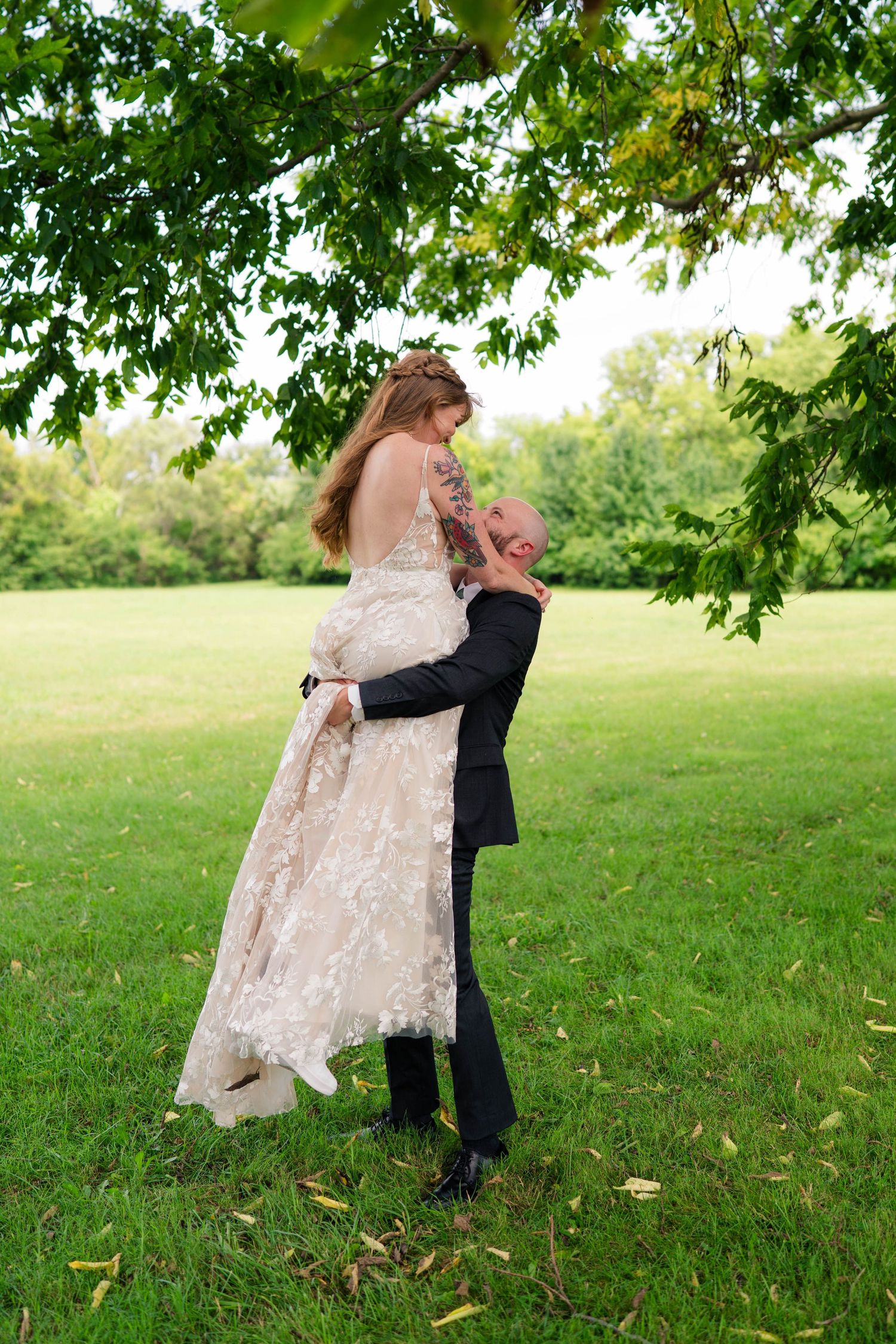 Groom lifting bride in lace wedding dress under green tree canopy in outdoor park setting.