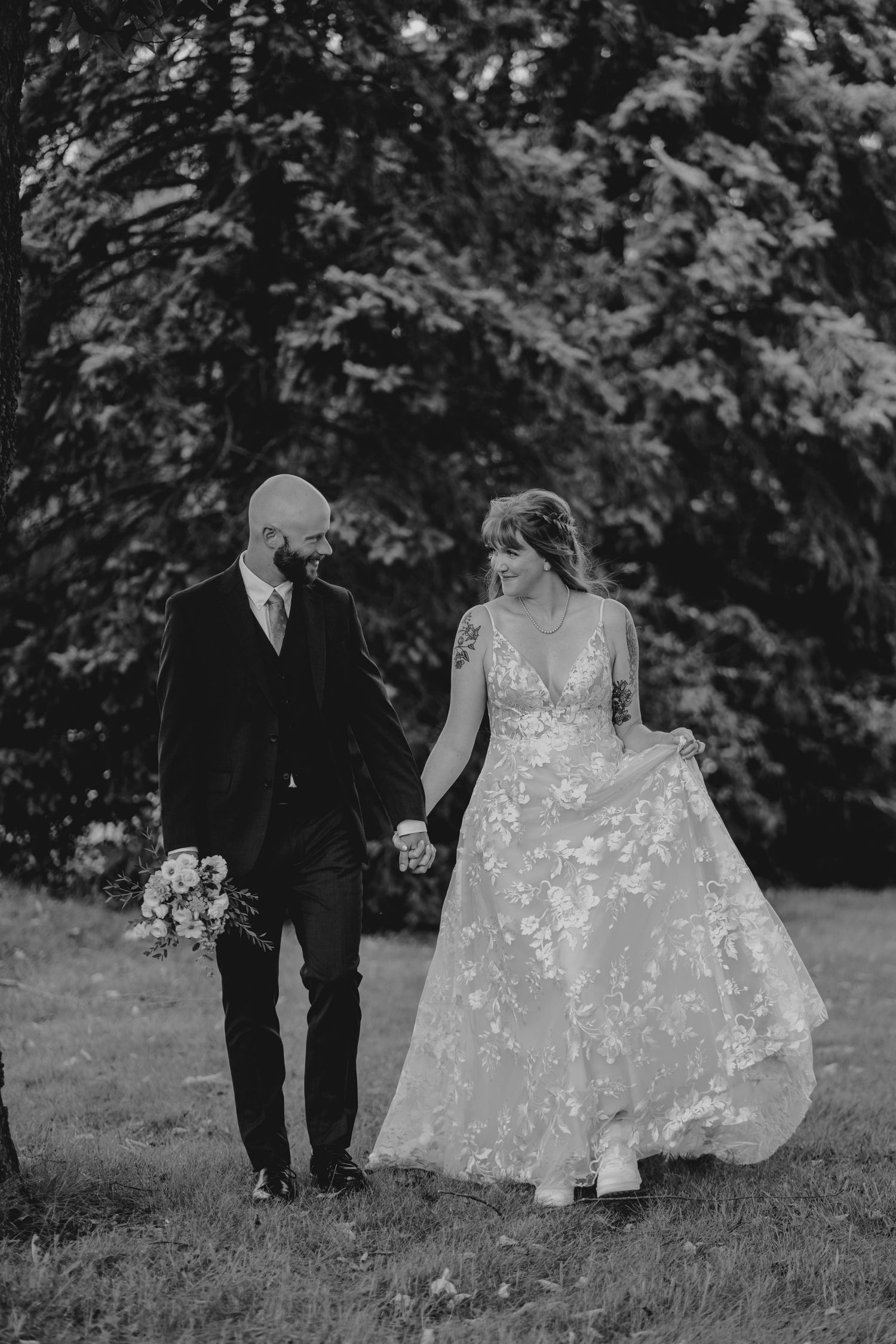 Black and white photo of a bride and groom walking hand in hand through a tree-lined outdoor setting.