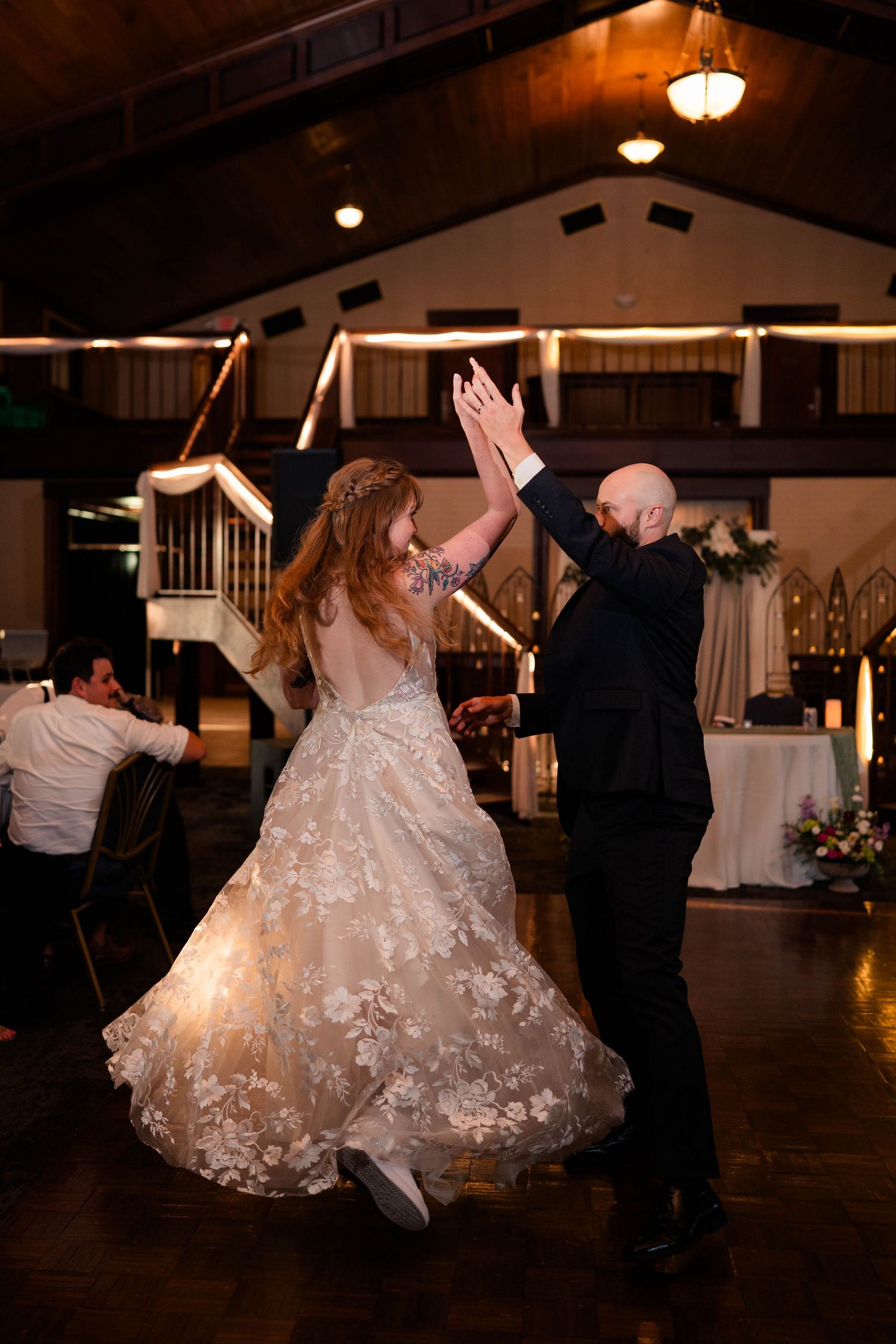 Bride in floral lace gown and groom high-five while dancing at elegant indoor wedding reception.