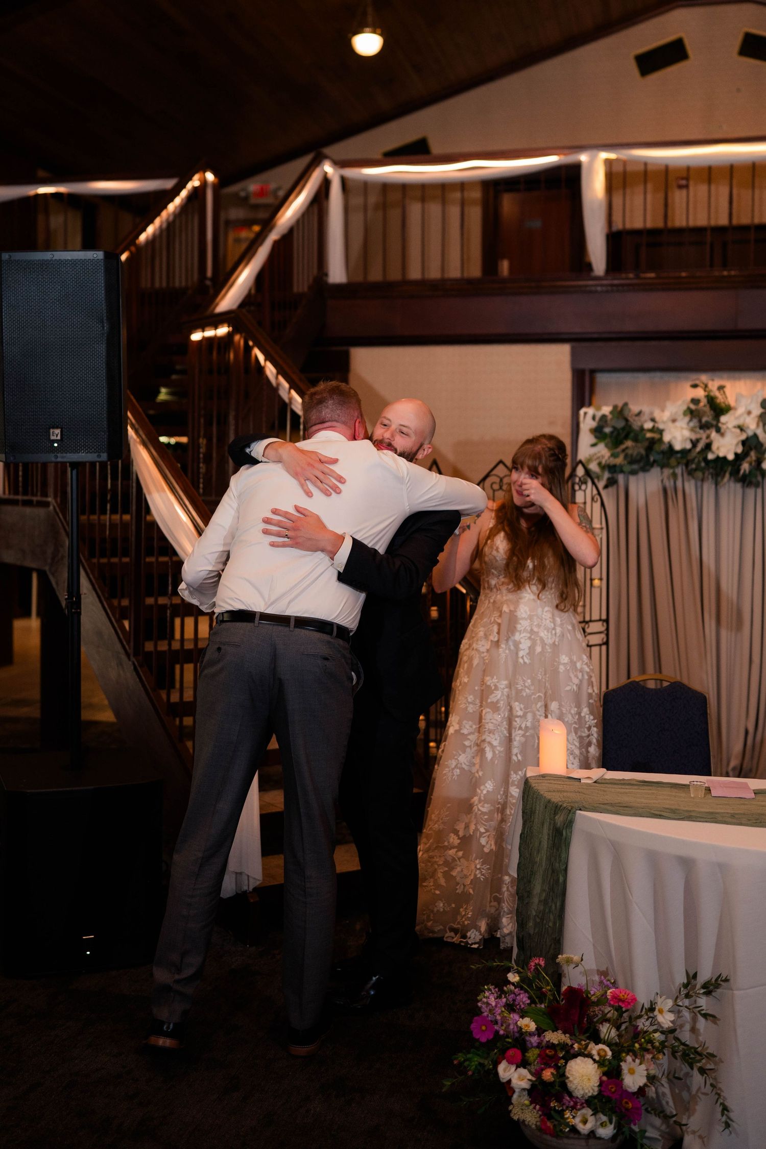 Groom embraces emotional guest near sweetheart table while bride watches at indoor wedding reception.