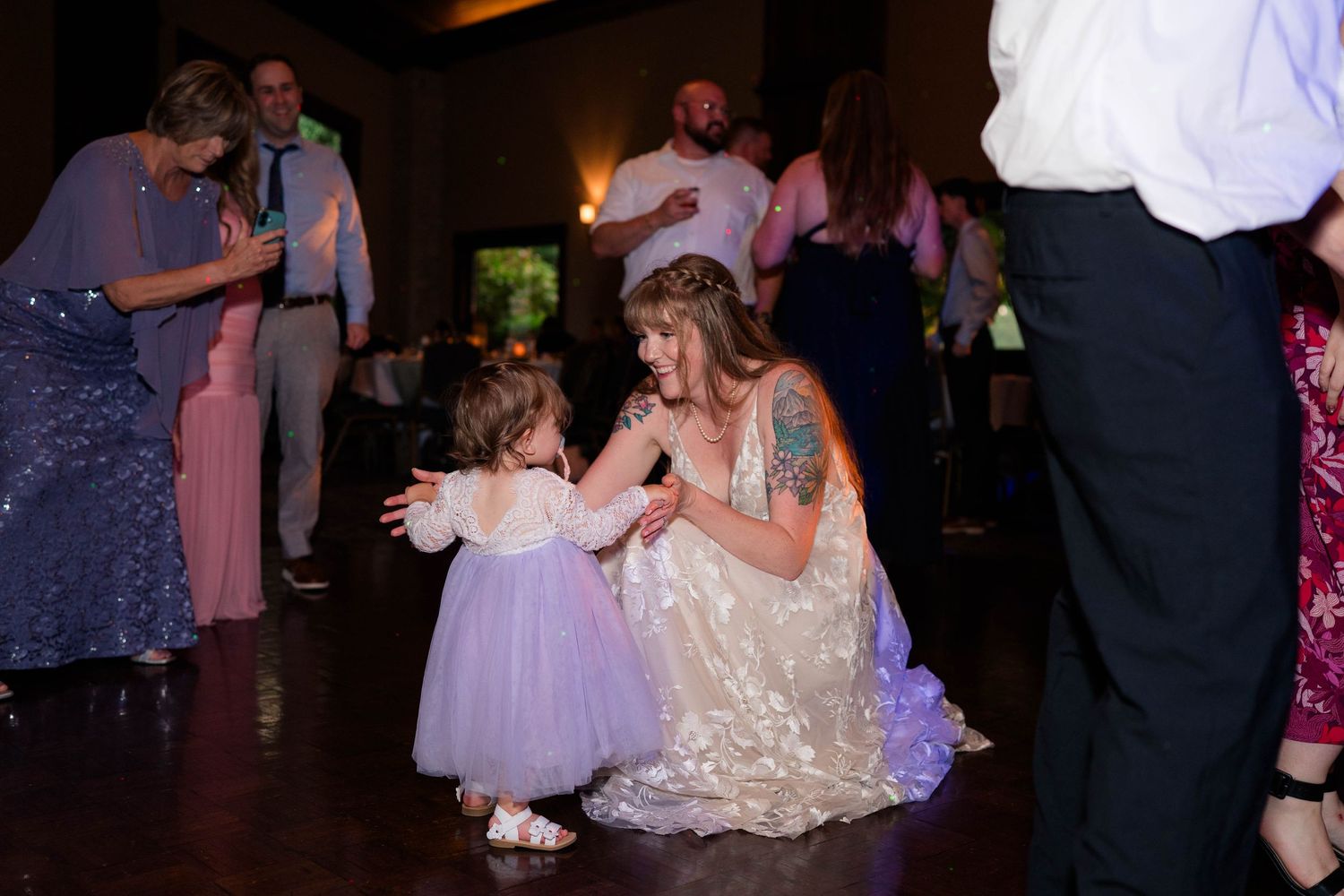 Bride crouches to interact with a little flower girl in lavender dress on dance floor.