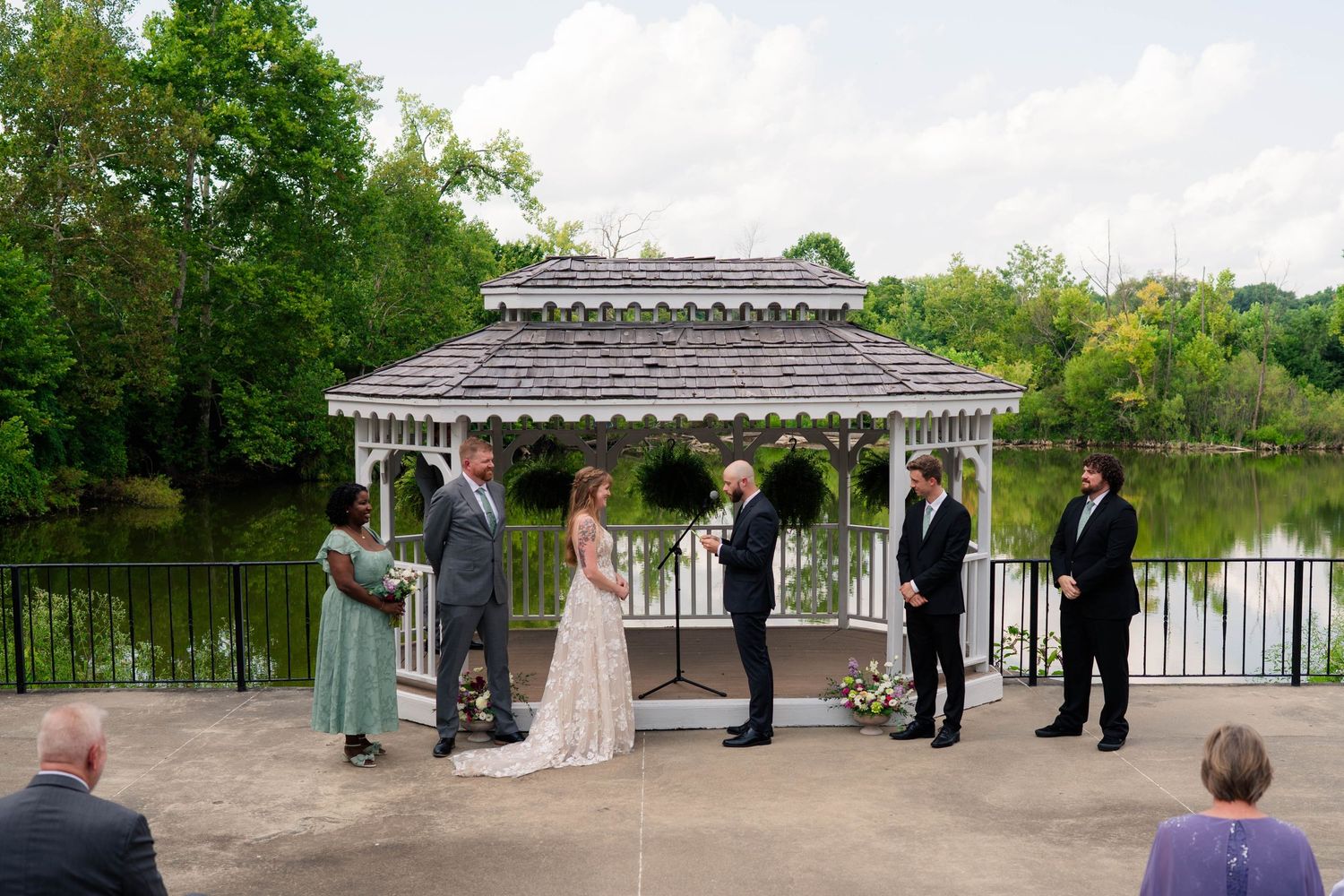 Outdoor wedding ceremony at a white gazebo beside a pond, with the couple exchanging vows before their guests.