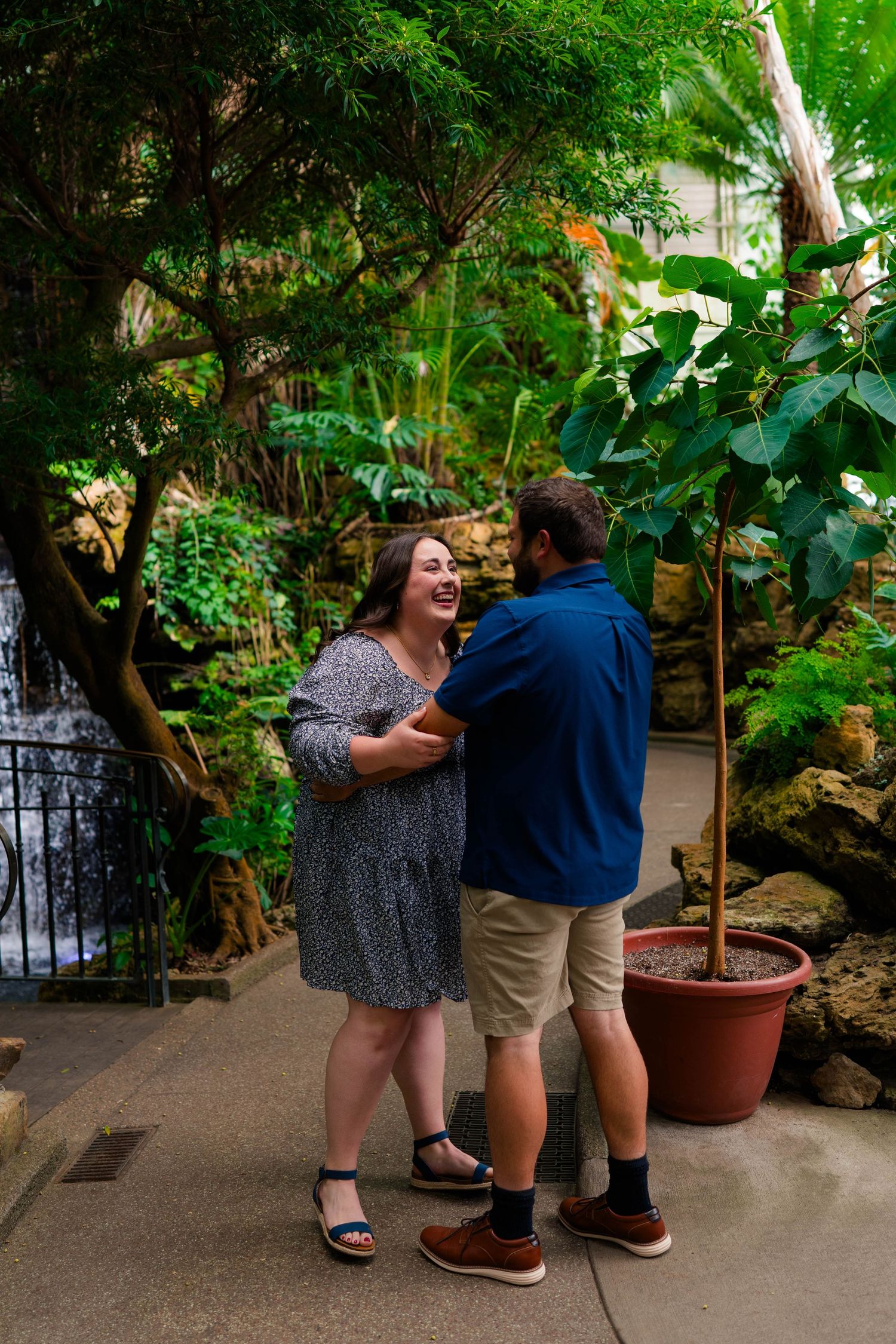 Newly engaged couple laughs together while holding hands near a waterfall in a tropical greenhouse.