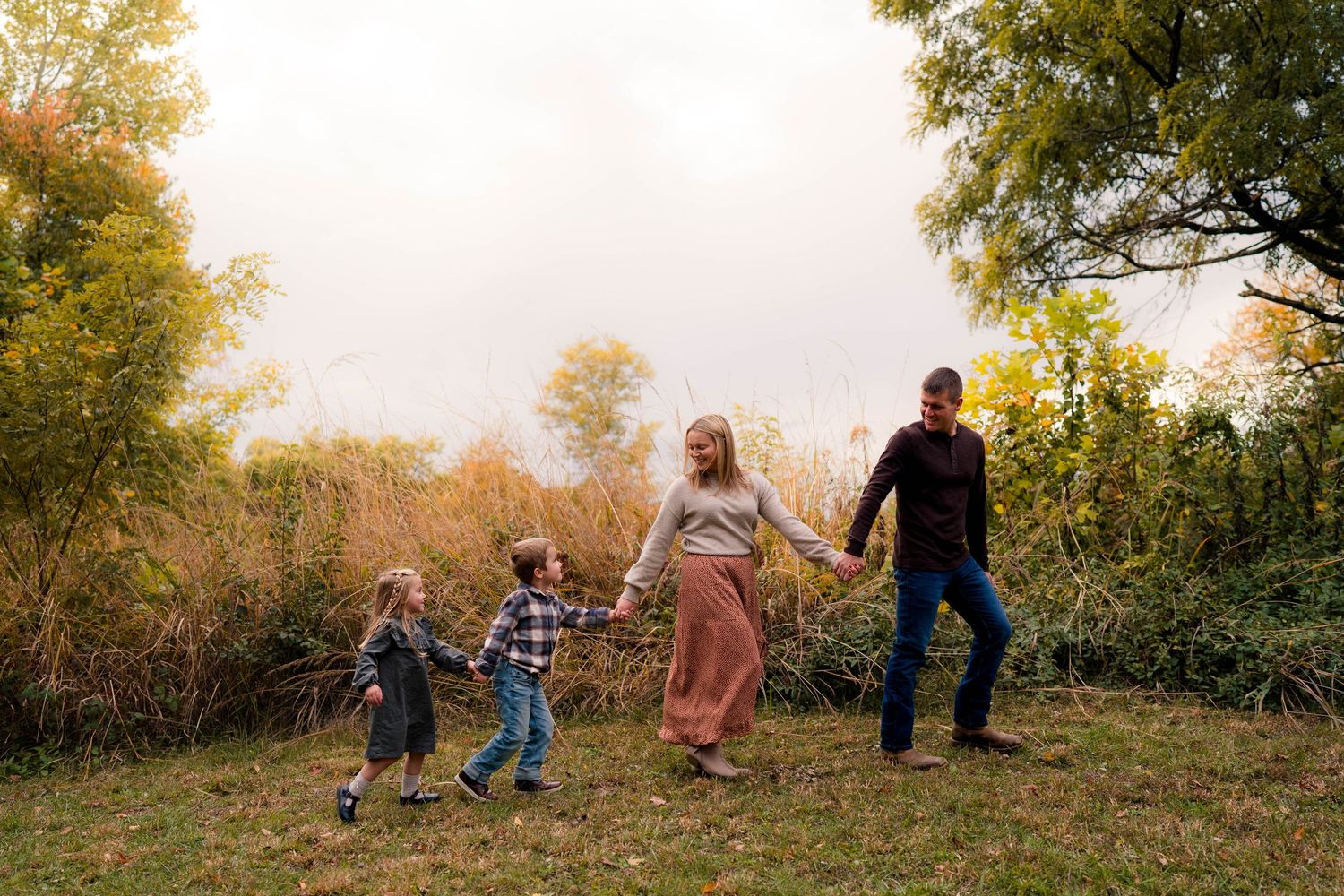Family of four holding hands walking through autumn meadow with colorful fall foliage.