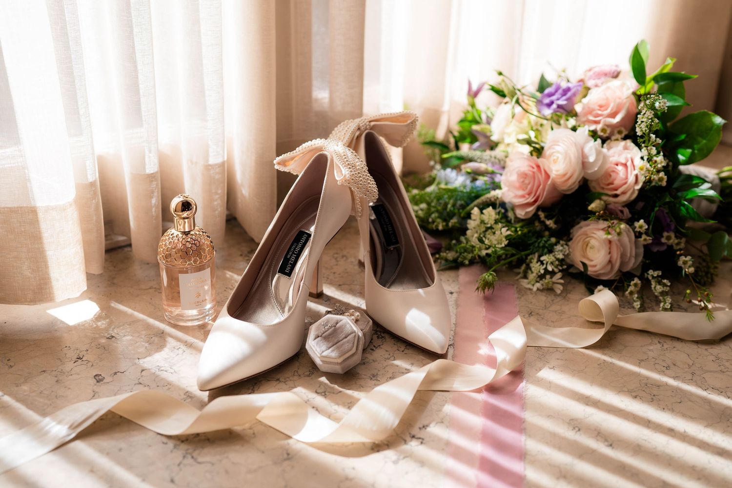 Elegant bridal accessories including white heels, a floral bouquet, and perfume arranged in soft natural window light.