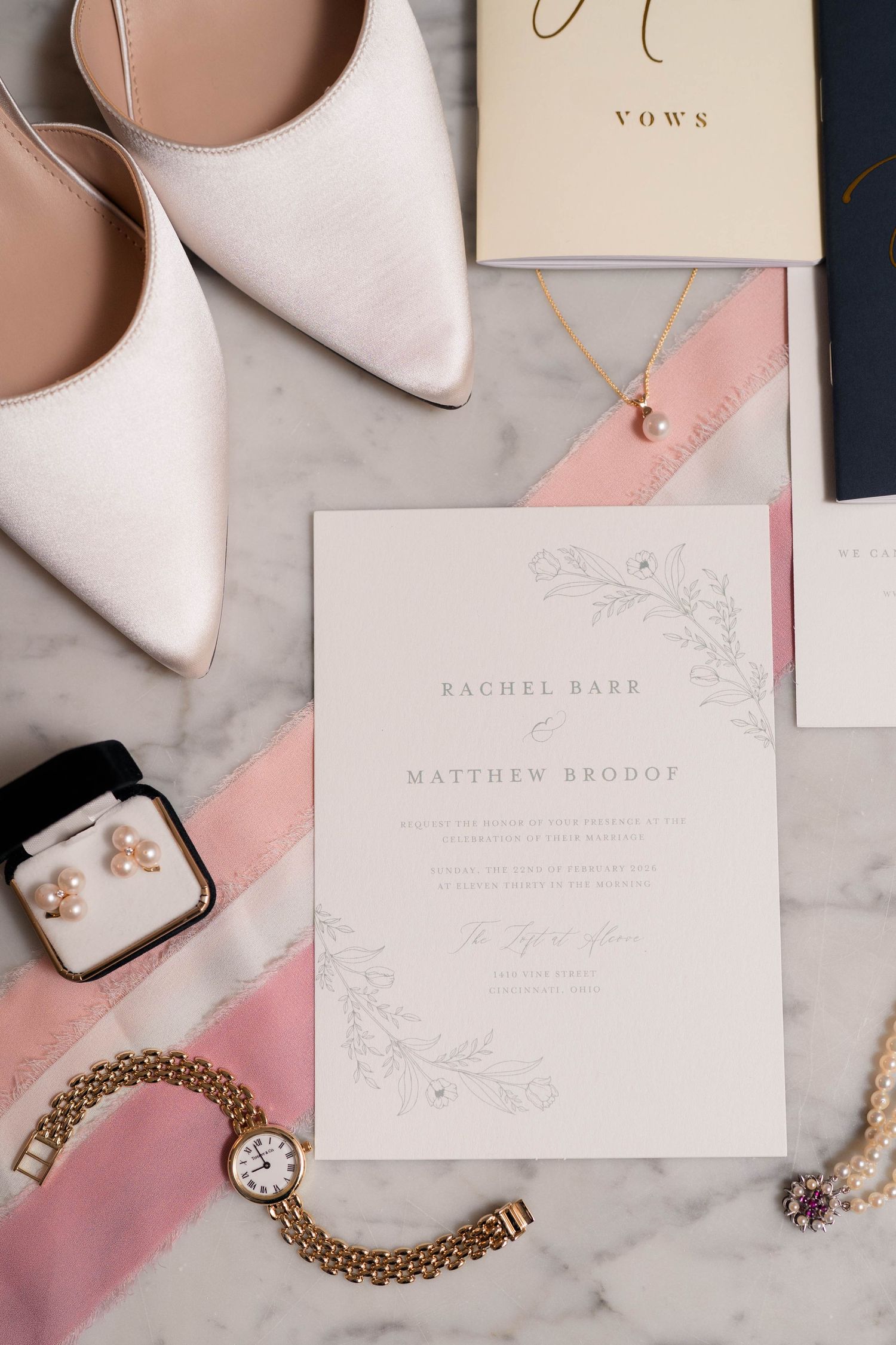 Flat lay of wedding invitation, white heels, pearl necklace, earrings, and gold watch on marble.