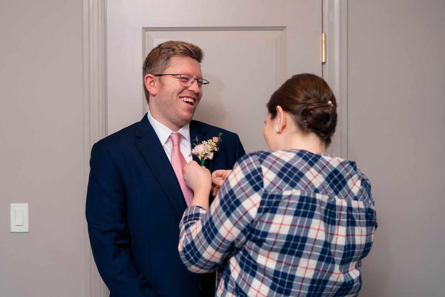 Laughing groom in navy suit and pink tie having a boutonniere pinned to his lapel.