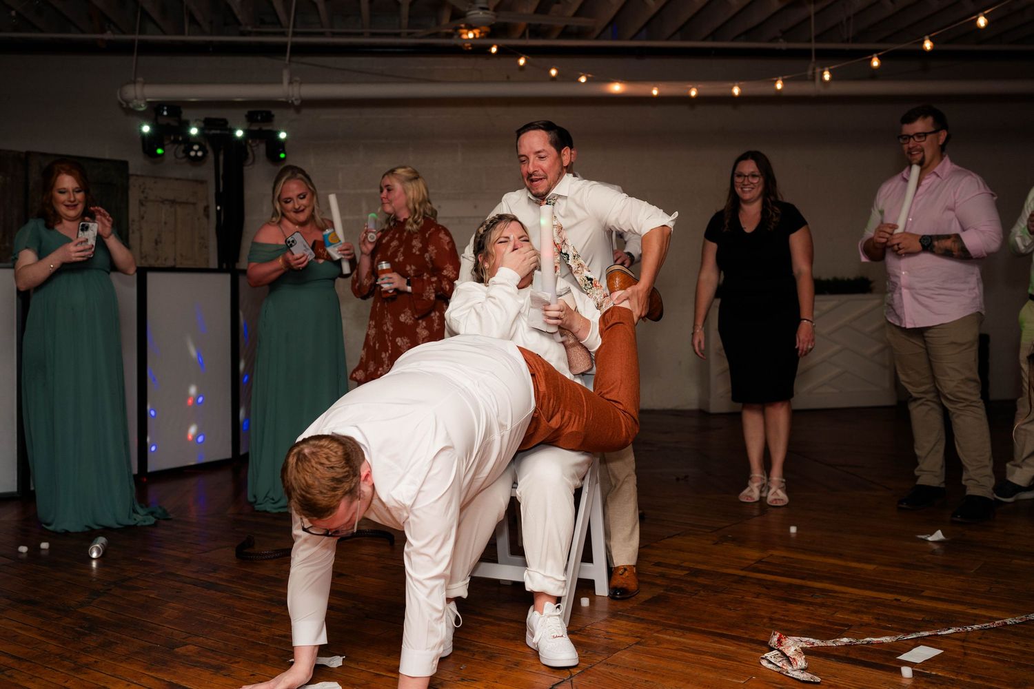 Groom retrieving garter from bride at lively wedding reception with guests watching and cheering.