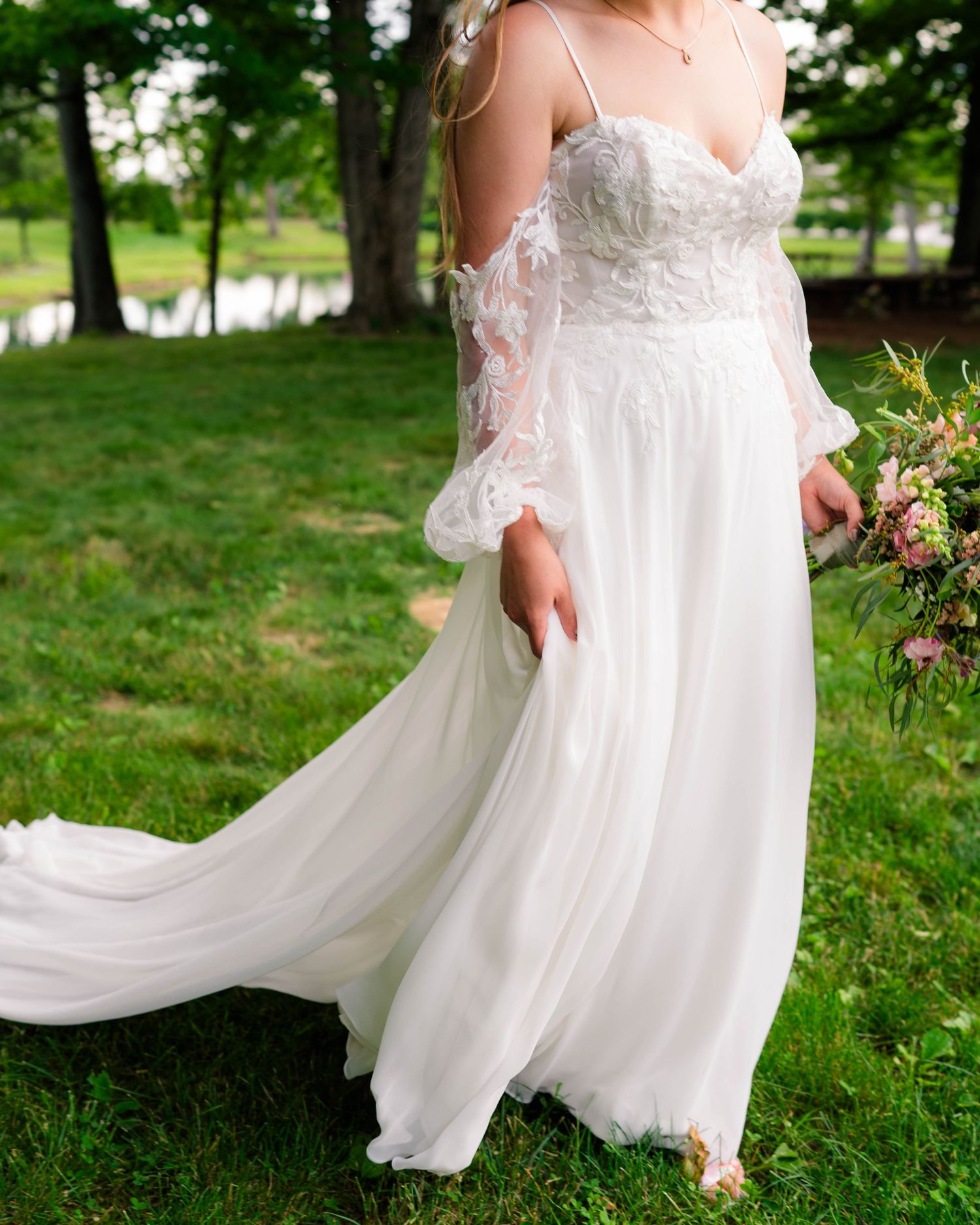Bride in flowing white lace off-shoulder wedding gown holding bouquet outdoors in lush greenery.