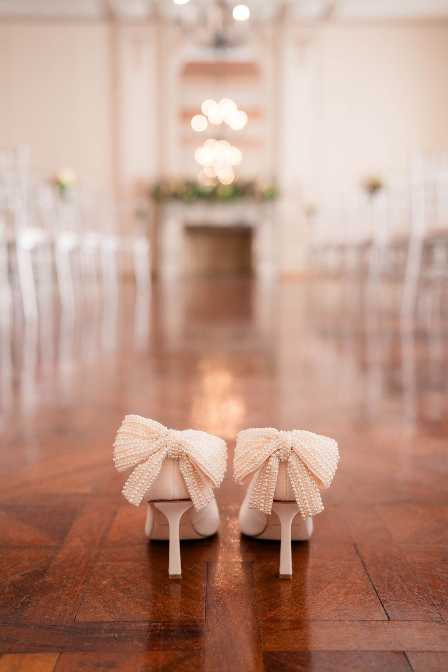 White bridal heels with pearl bow detail on hardwood floor with blurred ceremony backdrop.
