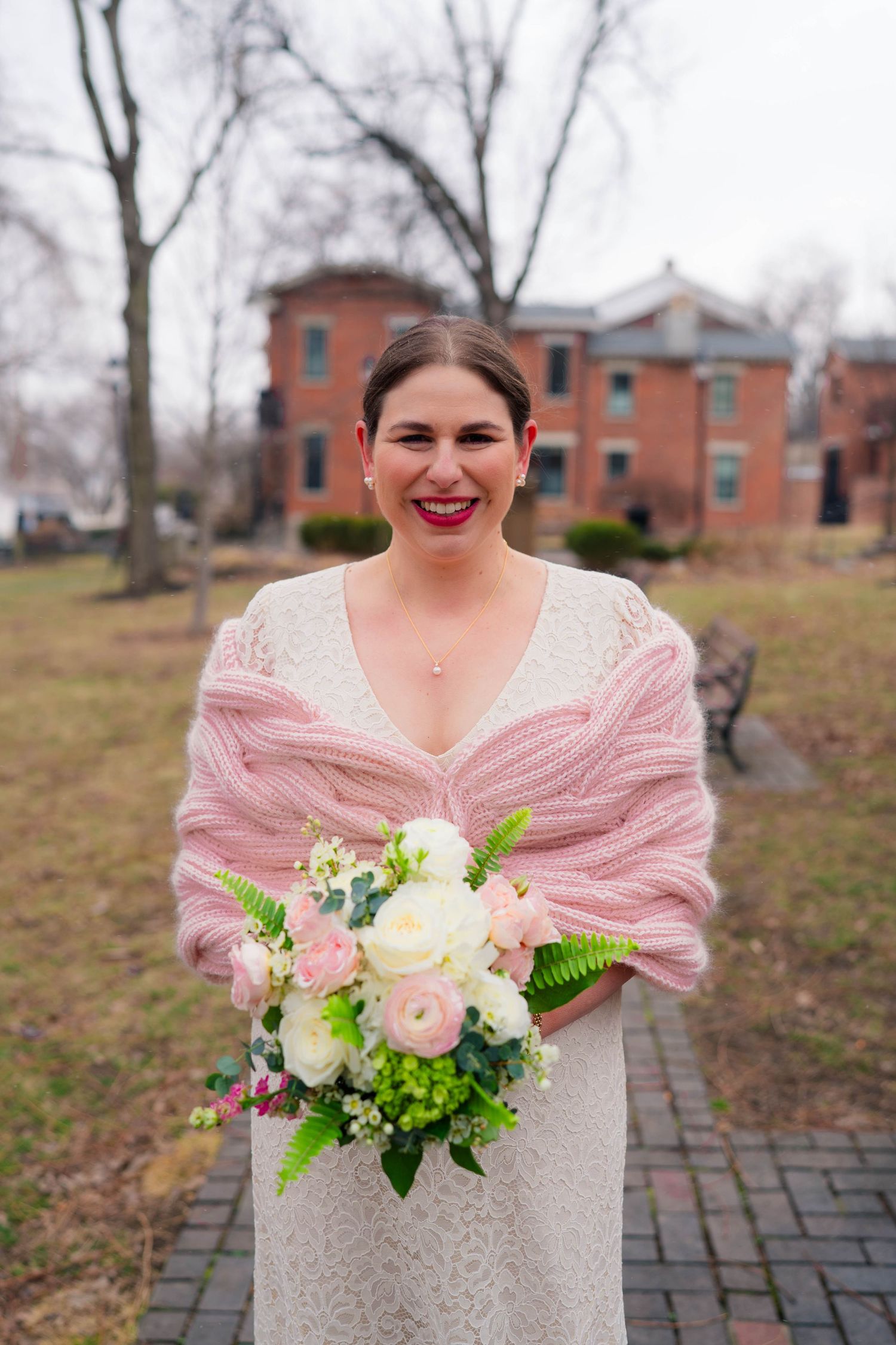 Smiling bride with red lips in lace gown and pink shawl holding a pink and white bouquet outdoors in winter.