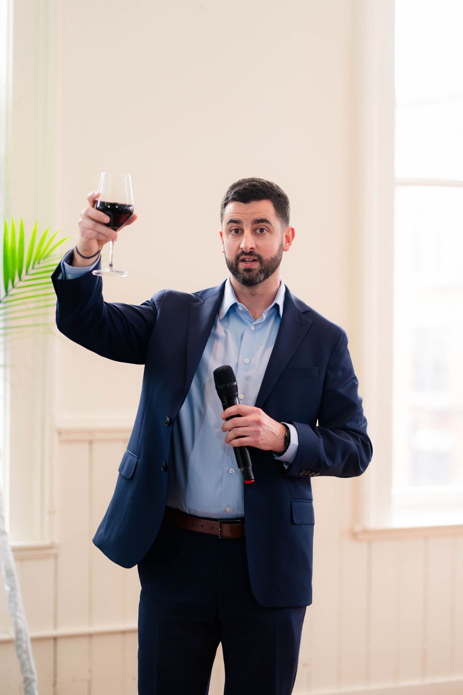 Man in navy suit raising a glass of red wine while holding a microphone during a wedding toast.