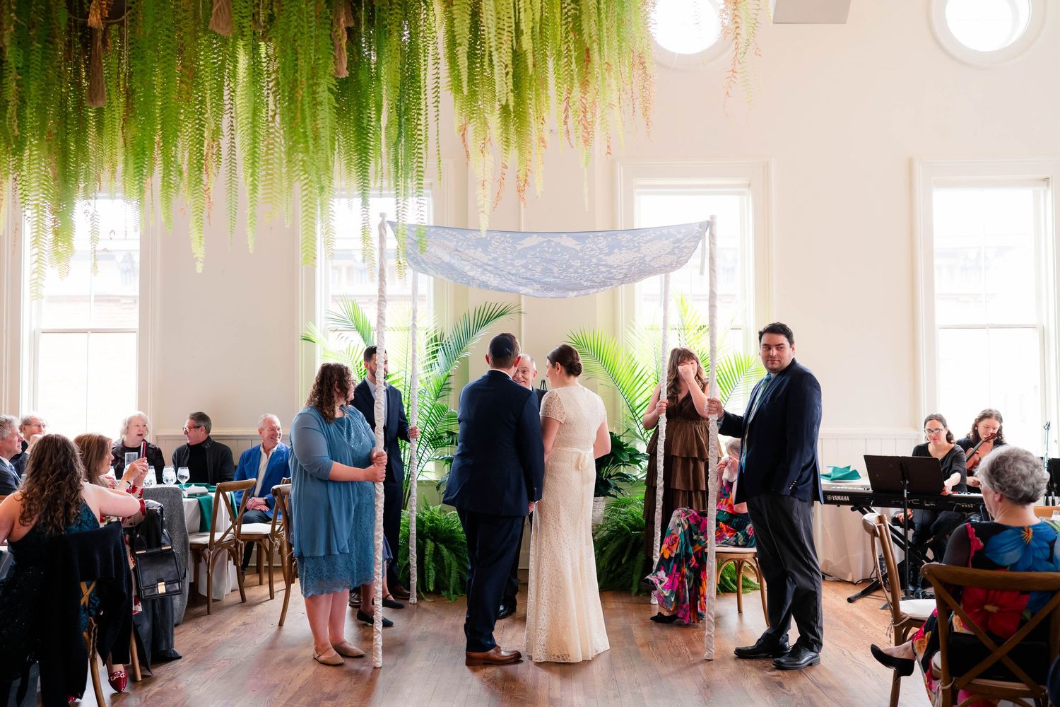 Wedding ceremony under hanging ferns and blue chuppah with officiant and wedding party gathered.