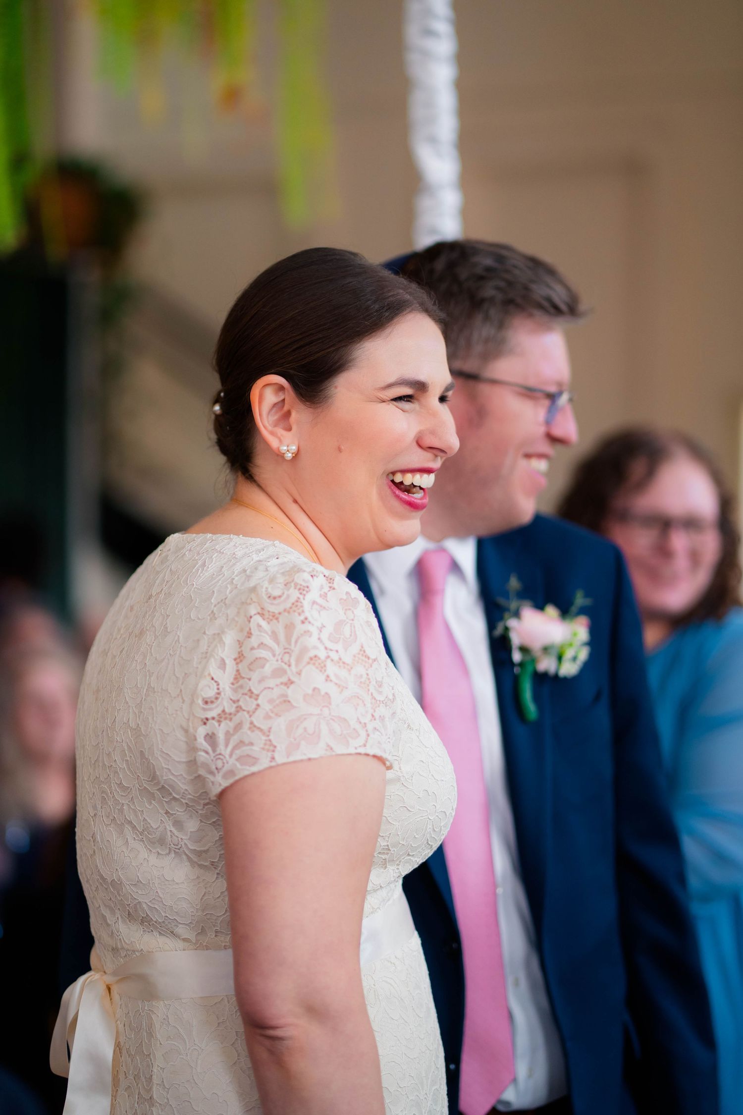 Laughing bride in lace gown stands beside groom in navy suit with pink tie at wedding.