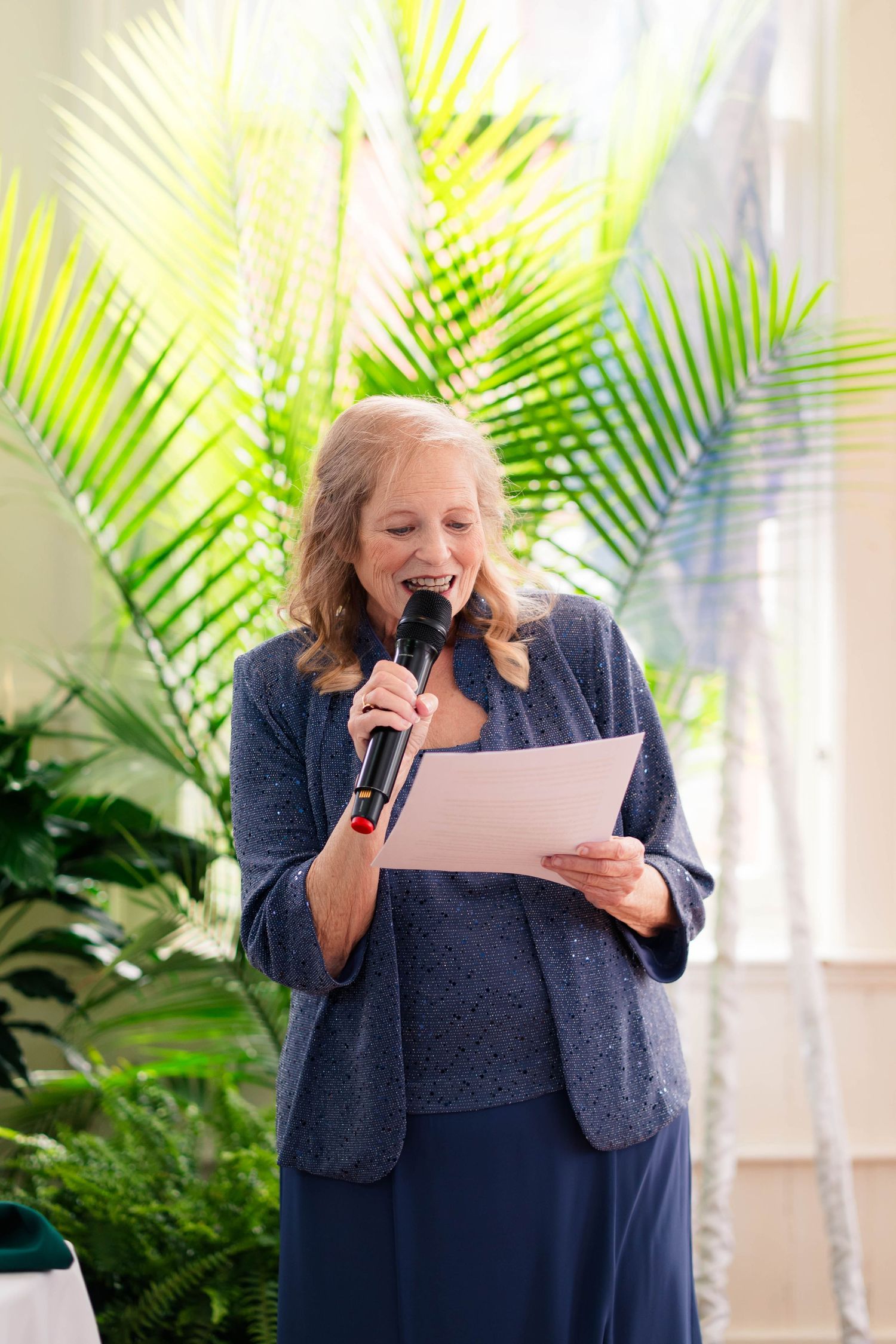 Smiling woman in blue dress delivers speech at wedding with tropical palm backdrop.