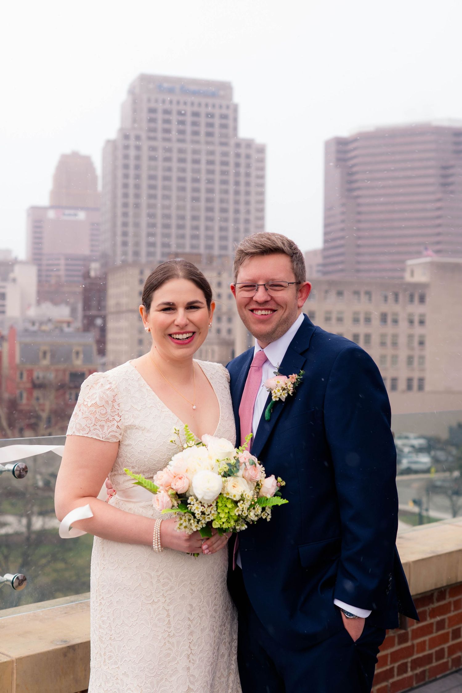 Happy bride holding pink and white bouquet with groom in navy suit and pink tie against city skyline.