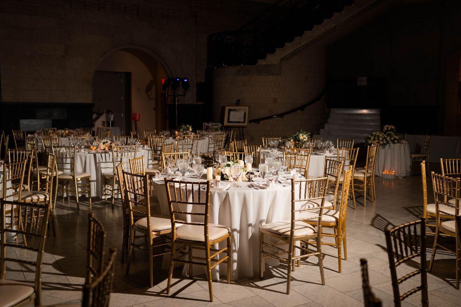 Elegant wedding reception hall with round tables, white linens, and gold chiavari chairs in dimly lit venue.