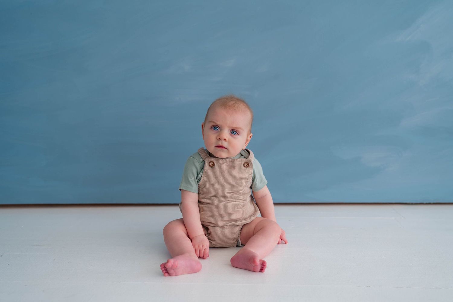 A cute baby in a sage green onesie and pink socks sits on a white floor against a blue backdrop.