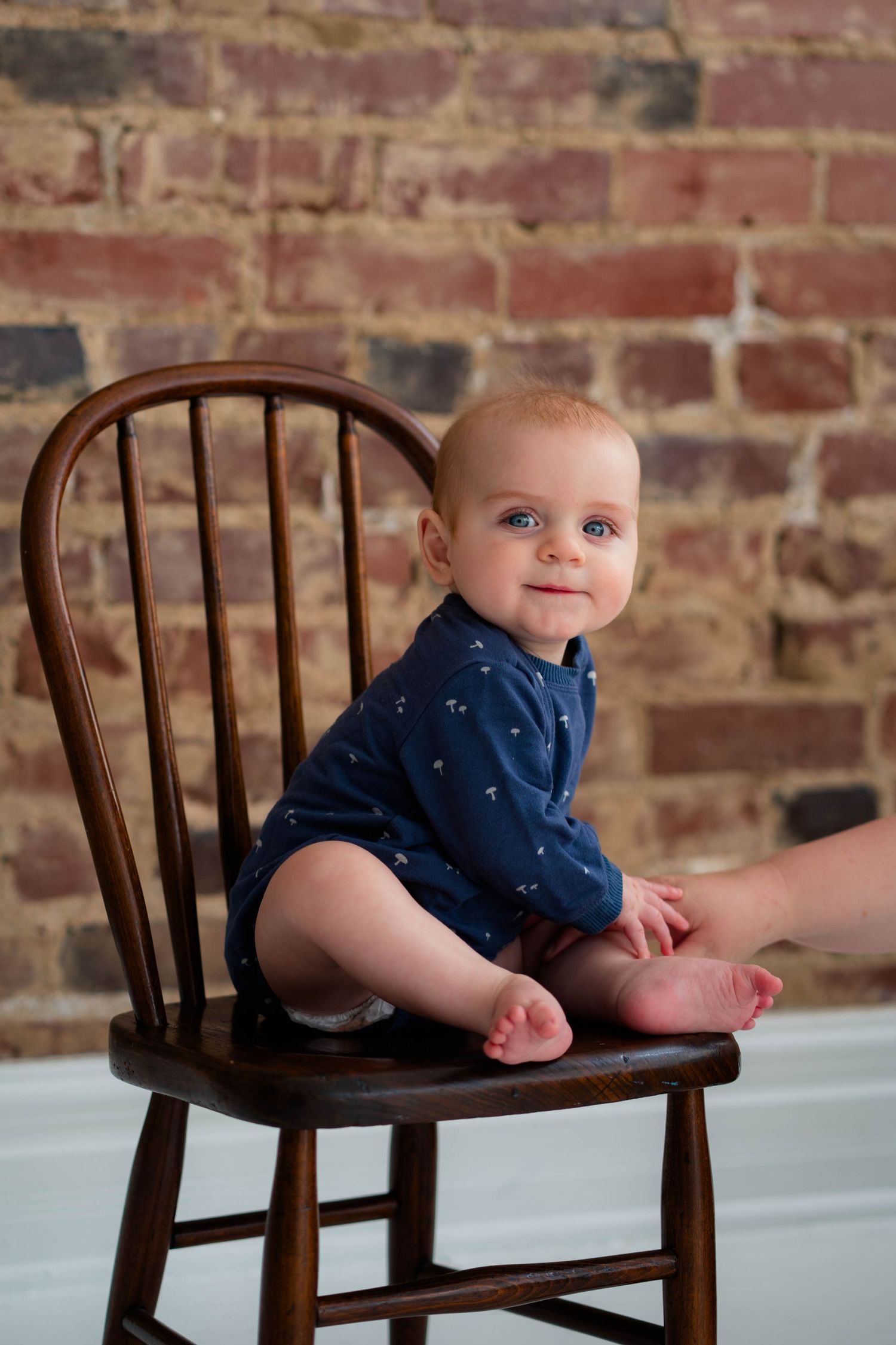 Smiling baby in navy romper sitting on wooden chair against rustic brick wall background.