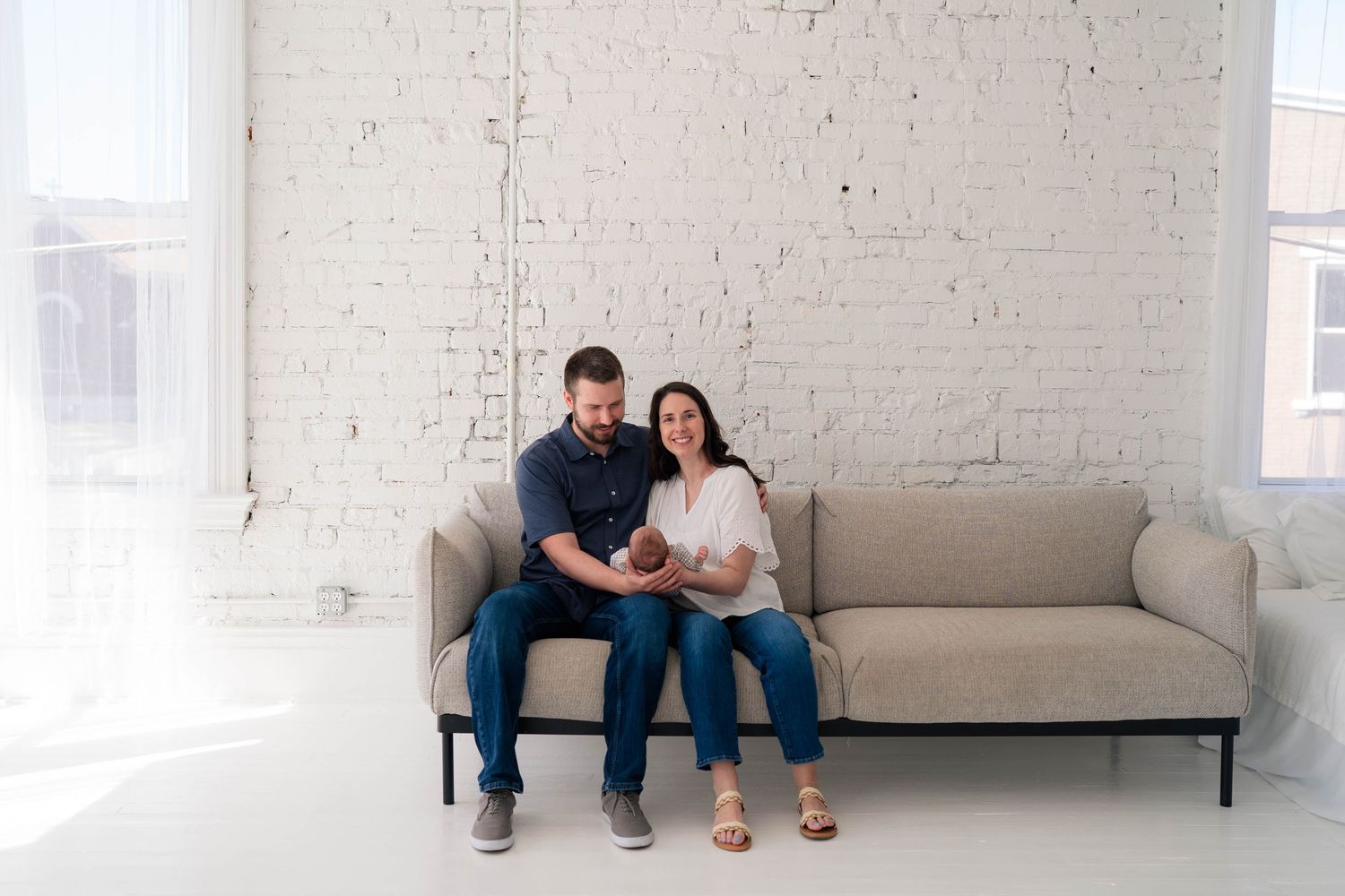 Happy couple sitting on a beige sofa holding their newborn baby in a bright white studio.