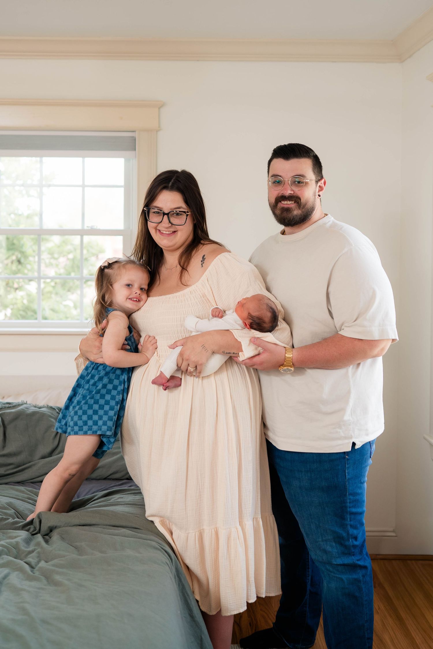 Happy family of four poses together with newborn baby and toddler daughter in a bright bedroom.