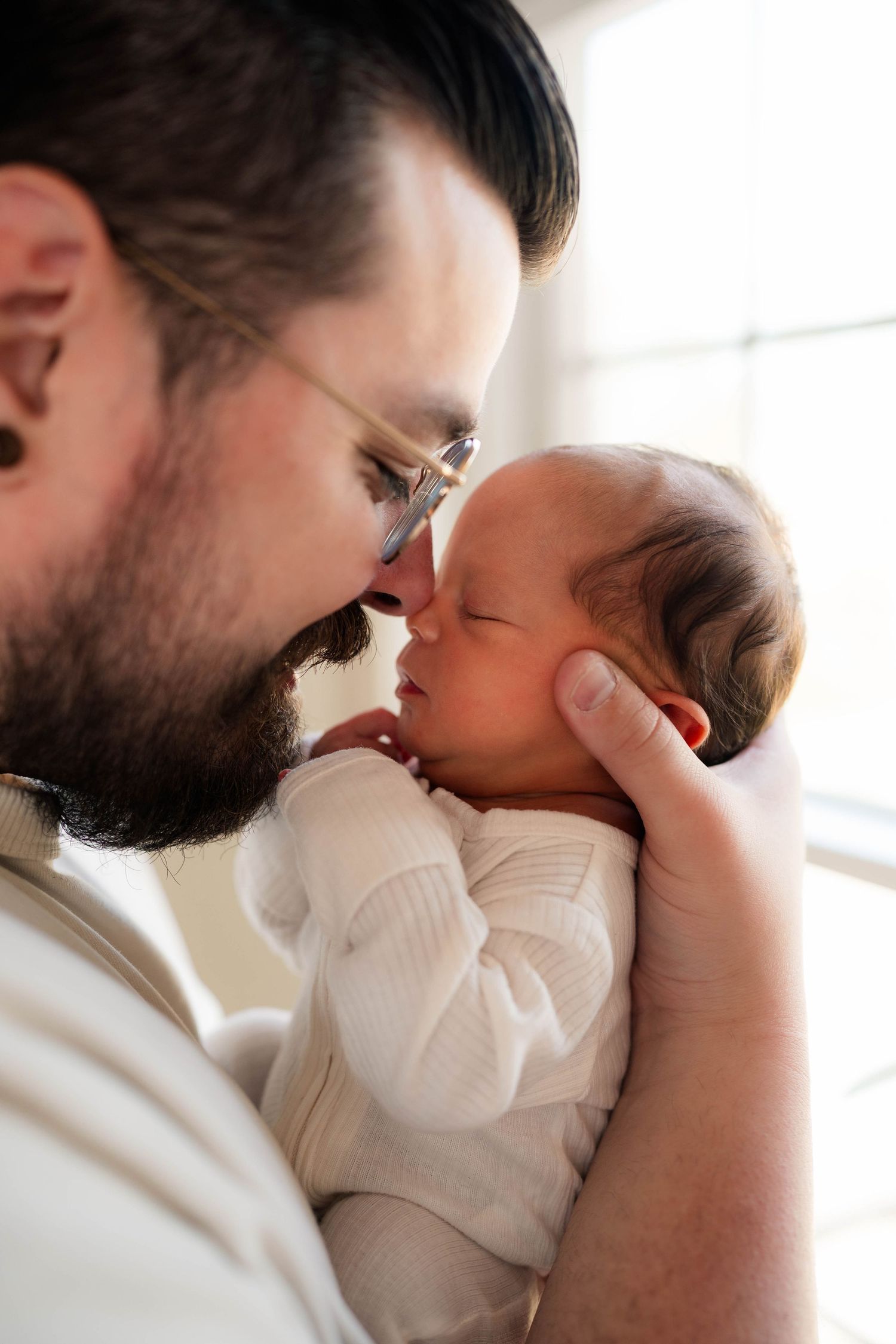 Bearded father in glasses tenderly touching foreheads with his sleeping newborn baby.