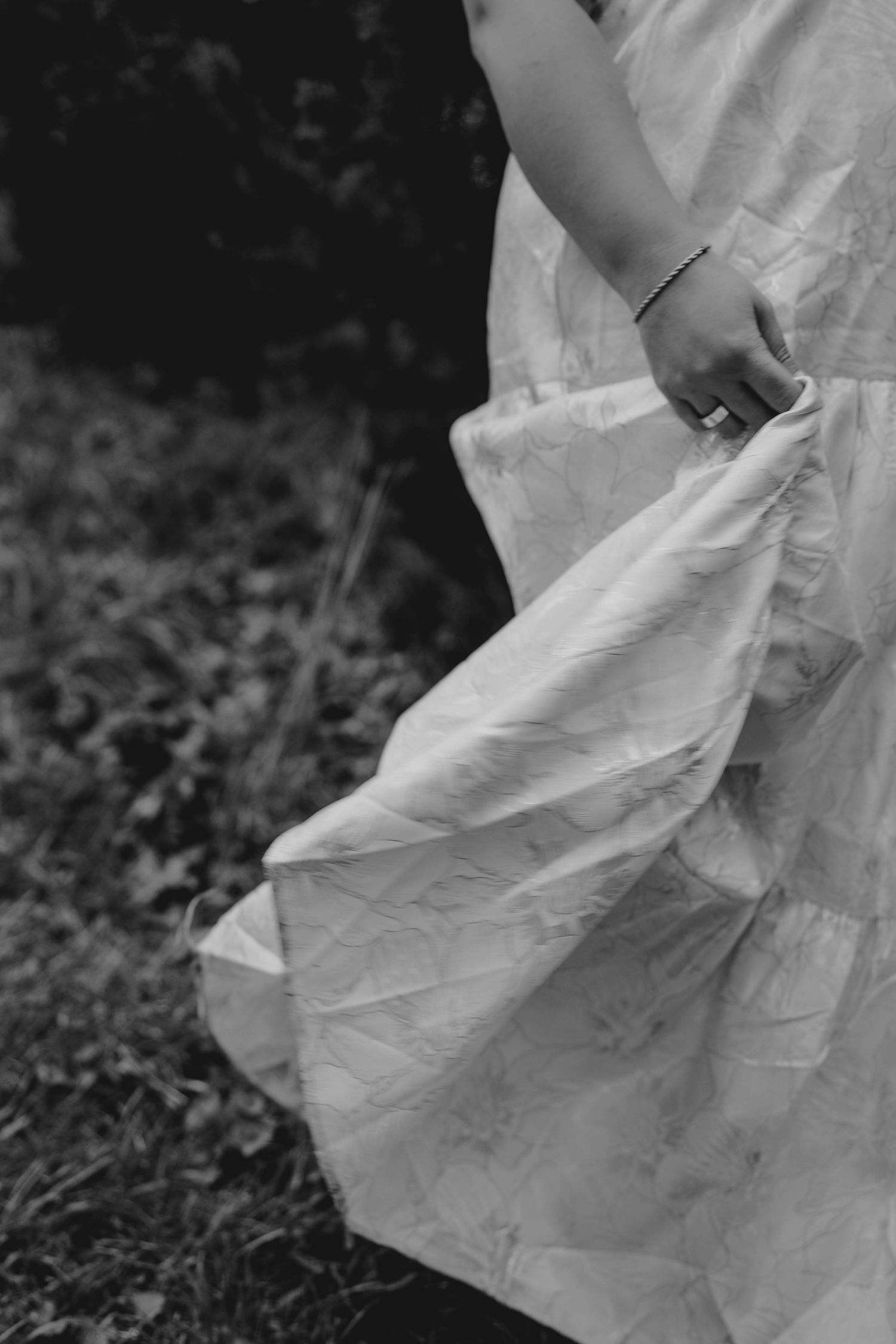 Black and white close-up of a bride holding her flowing white dress hem outdoors on grass.