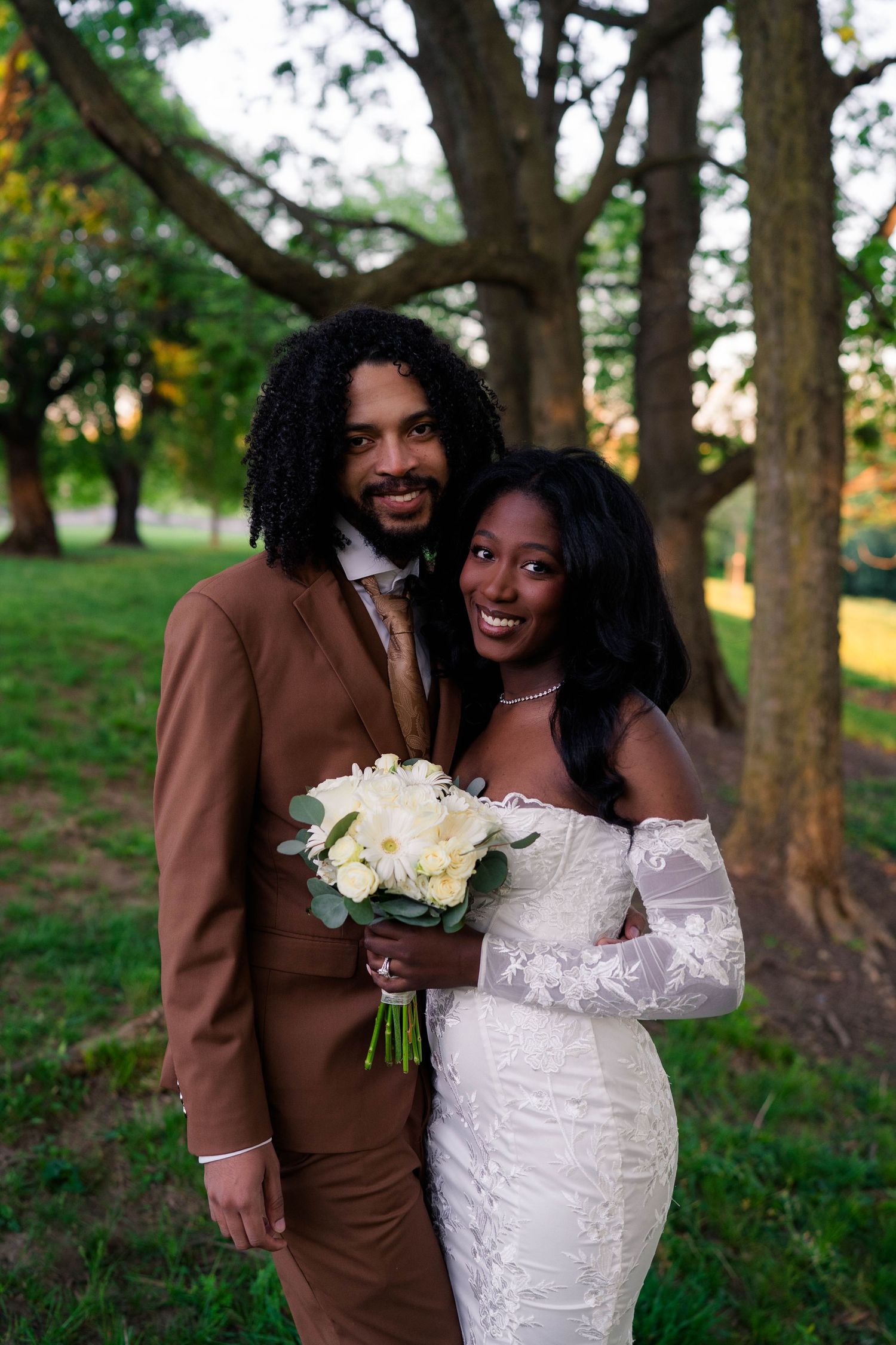 Smiling Black newlyweds pose outdoors, groom in brown suit, bride in off-shoulder lace gown holding white bouquet.