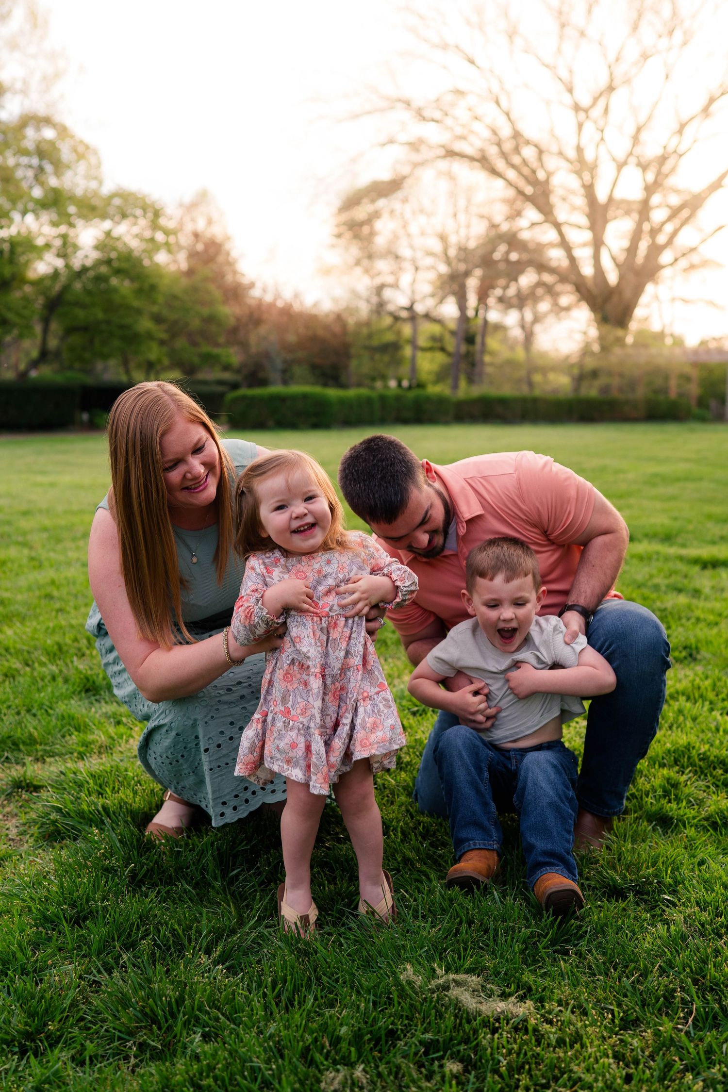 A happy family of four plays together on green grass at sunset, with parents and two young children laughing outdoors.