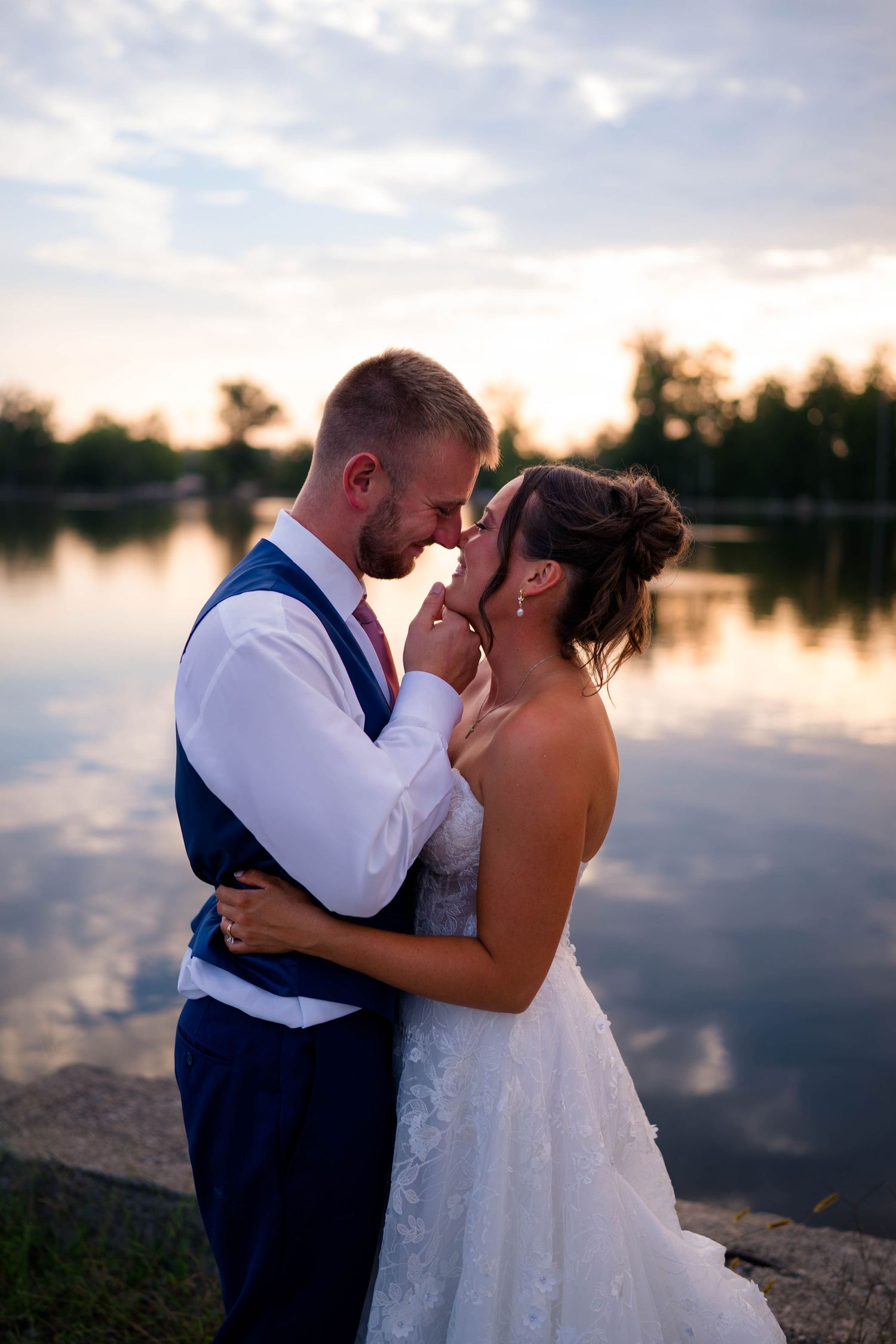 Bride and groom share a romantic forehead touch by a reflective lake at sunset, groom in navy vest, bride in lace gown.