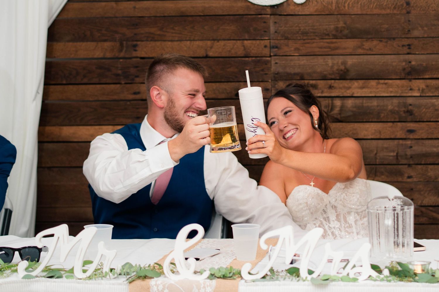 Bride and groom laugh while clinking drinks at their Mr. and Mrs. decorated wedding table.