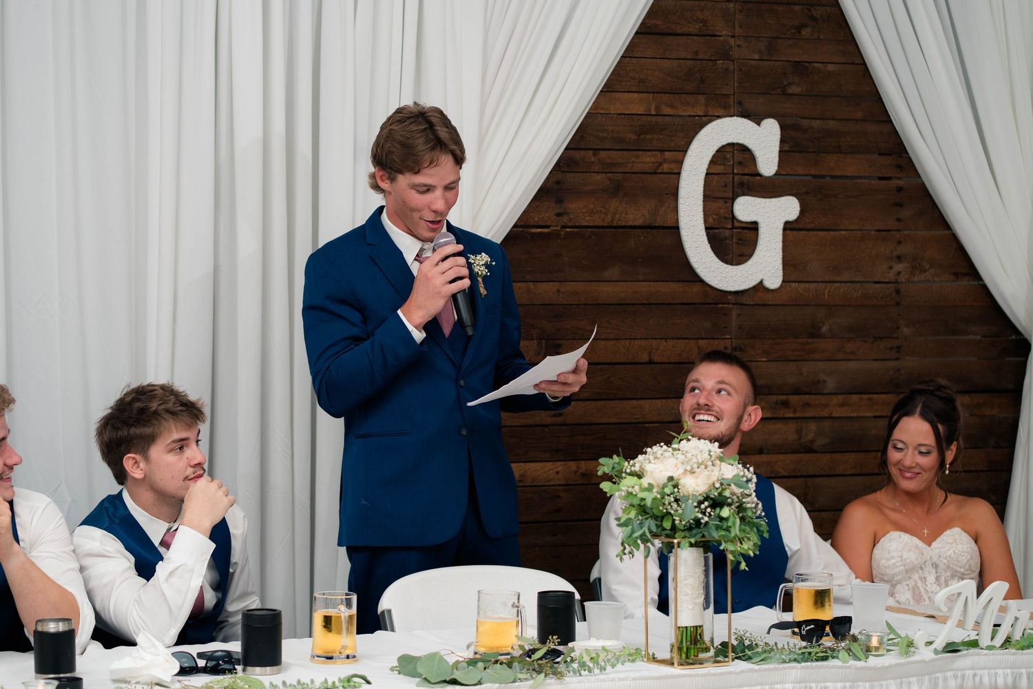 Best man in blue suit delivers speech at wedding reception as groom laughs at head table decorated with white flowers.
