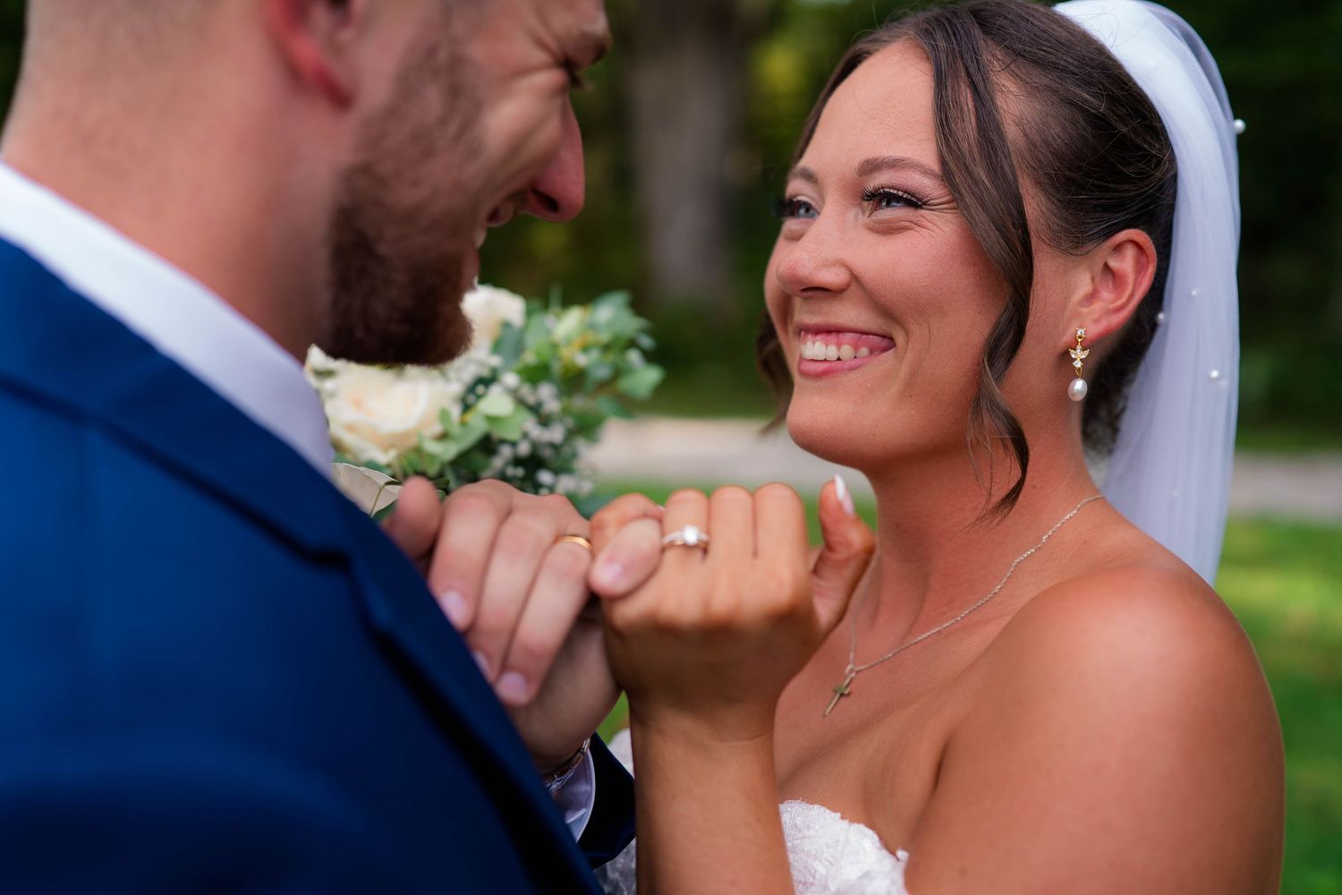Smiling bride shows off her wedding ring while groom holds her hand during outdoor portraits.