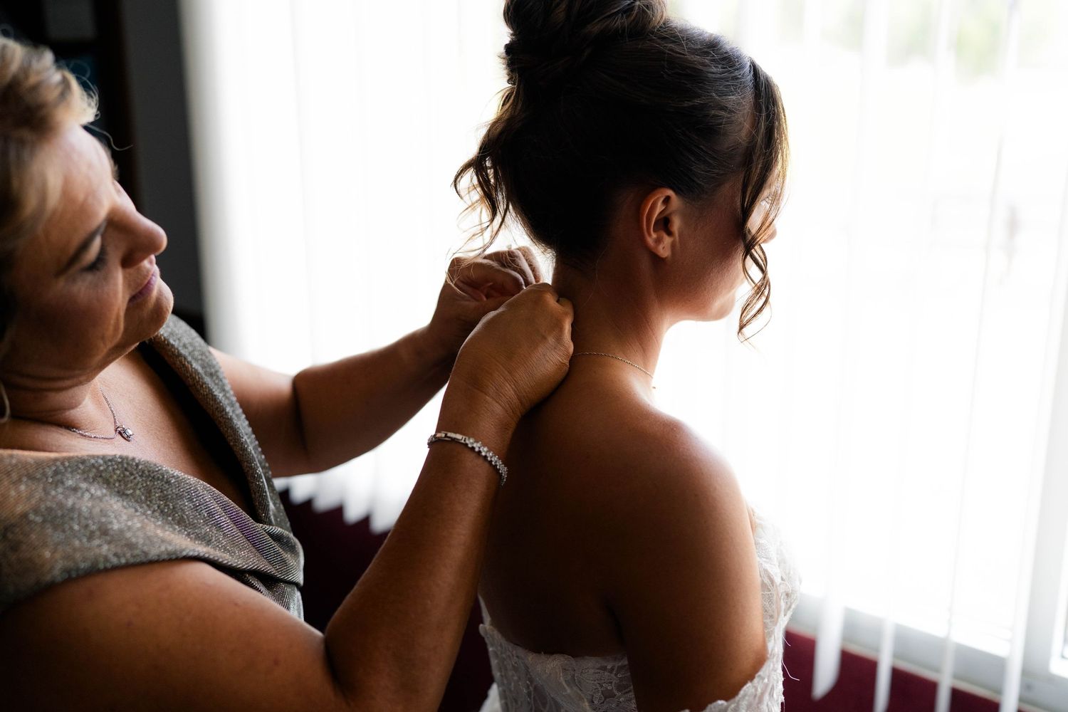 Older woman helping bride fasten necklace near window on wedding day.