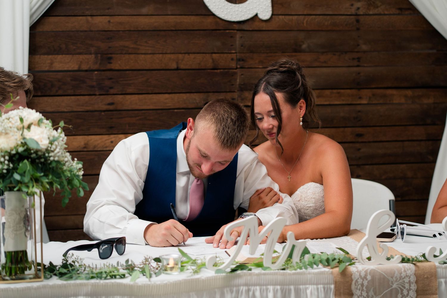 Newlyweds sign marriage certificate at decorated sweetheart table during wedding reception.